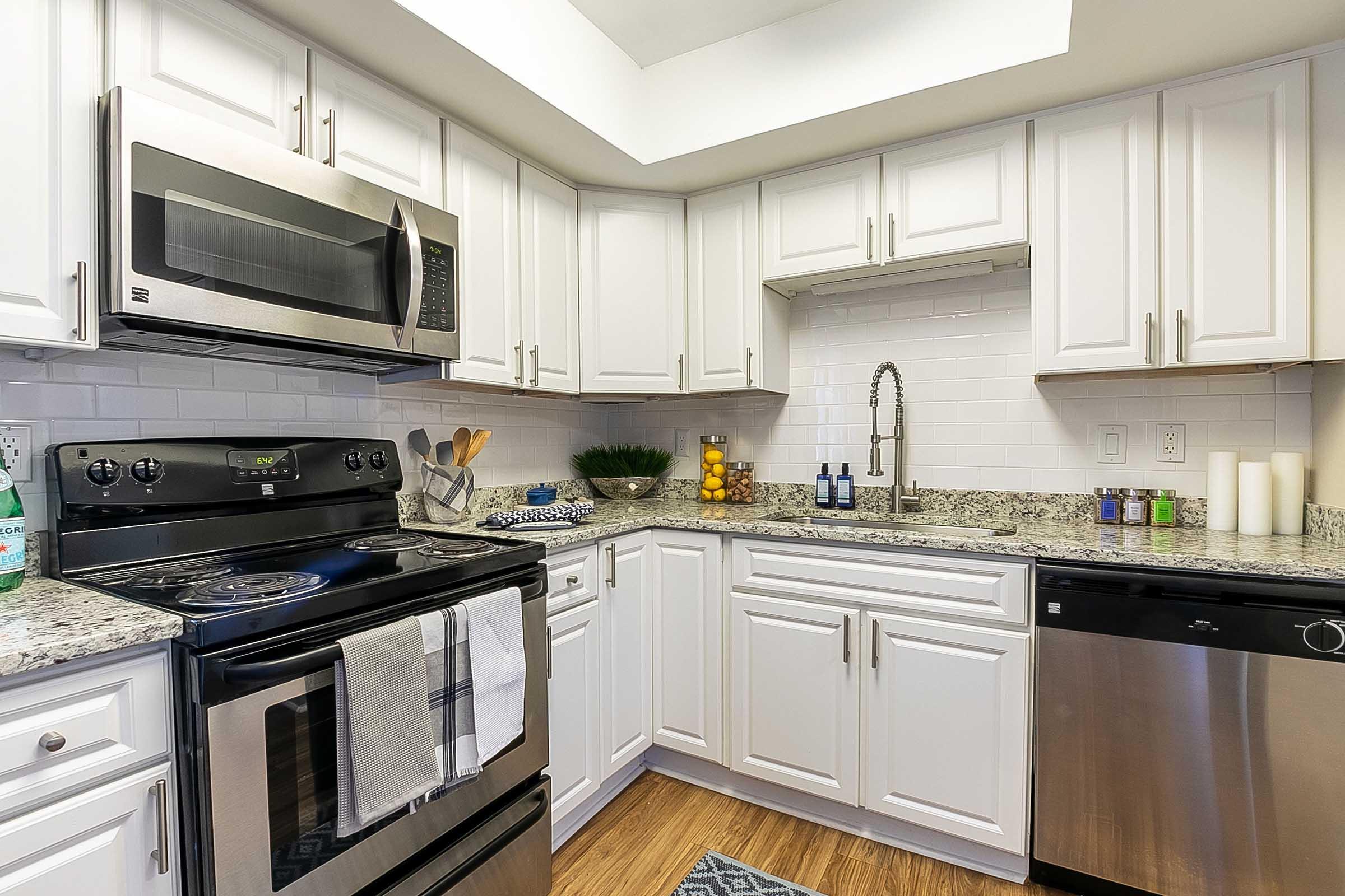 Modern kitchen featuring white cabinets, granite countertops, stainless steel appliances including a microwave, stove, and dishwasher. The space is well-lit with a light fixture and includes decorative items like a fruit bowl and kitchen utensils. Hardwood flooring adds warmth to the design.