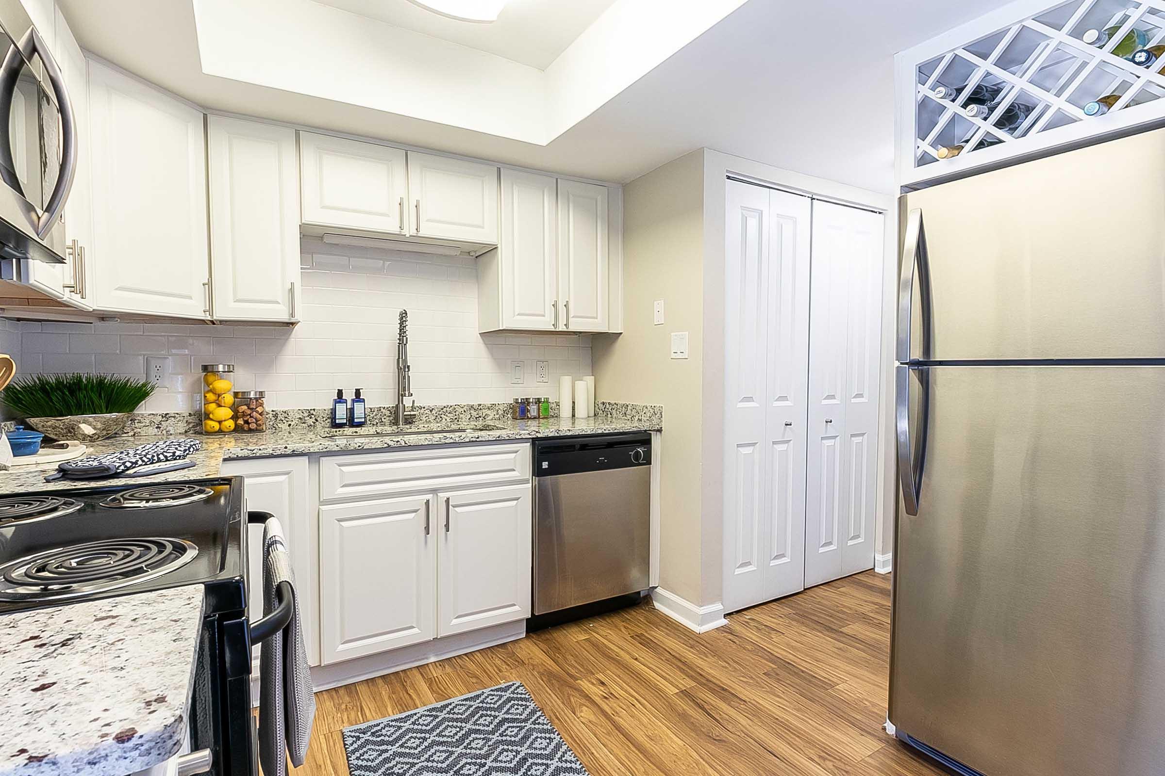 Modern kitchen featuring white cabinets, granite countertops, a stainless steel refrigerator, and a black stove. The space includes a dishwasher and decorative items like fruits on the countertop. Bright lighting enhances the clean, organized look of the kitchen.