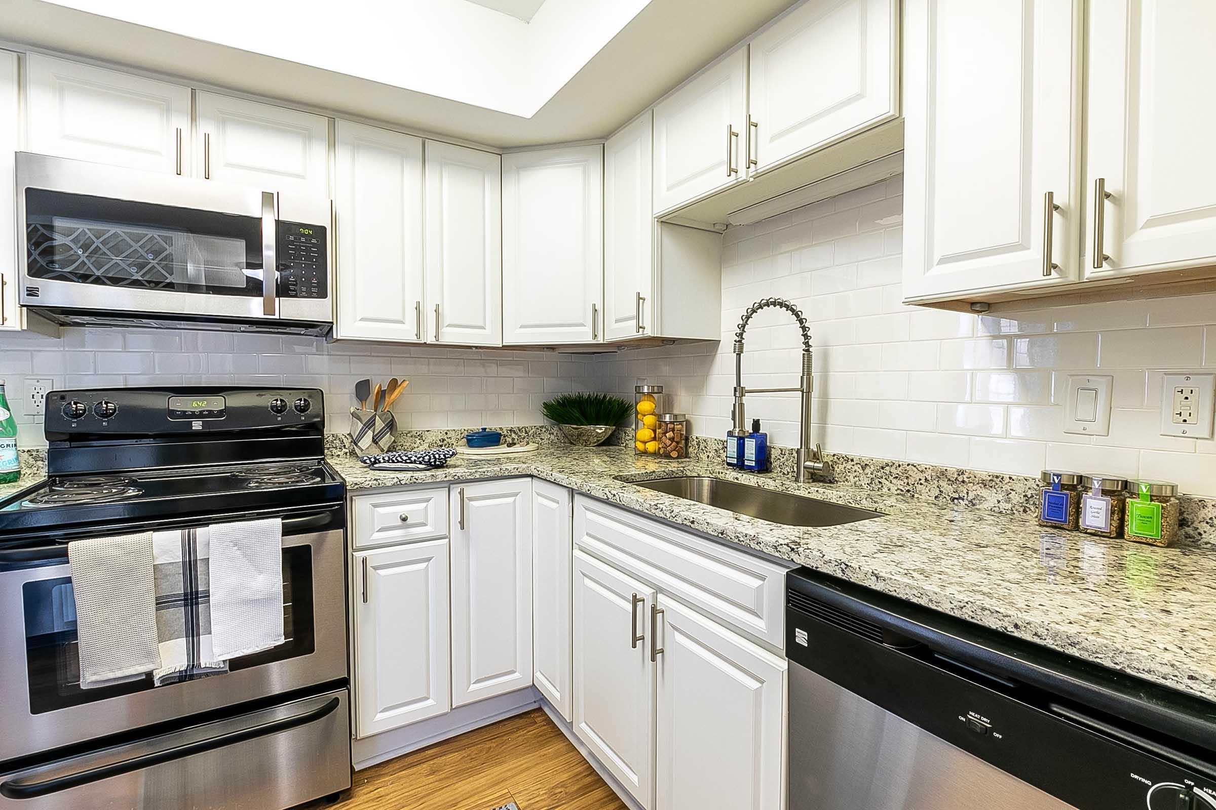 A modern kitchen featuring white cabinets, a stainless steel fridge, microwave, and stove, a granite countertop, and a stainless steel sink with a pull-down faucet. Decorative items include a bowl of lemons and colorful bottles on the counter. The floor is a light wood-like material.
