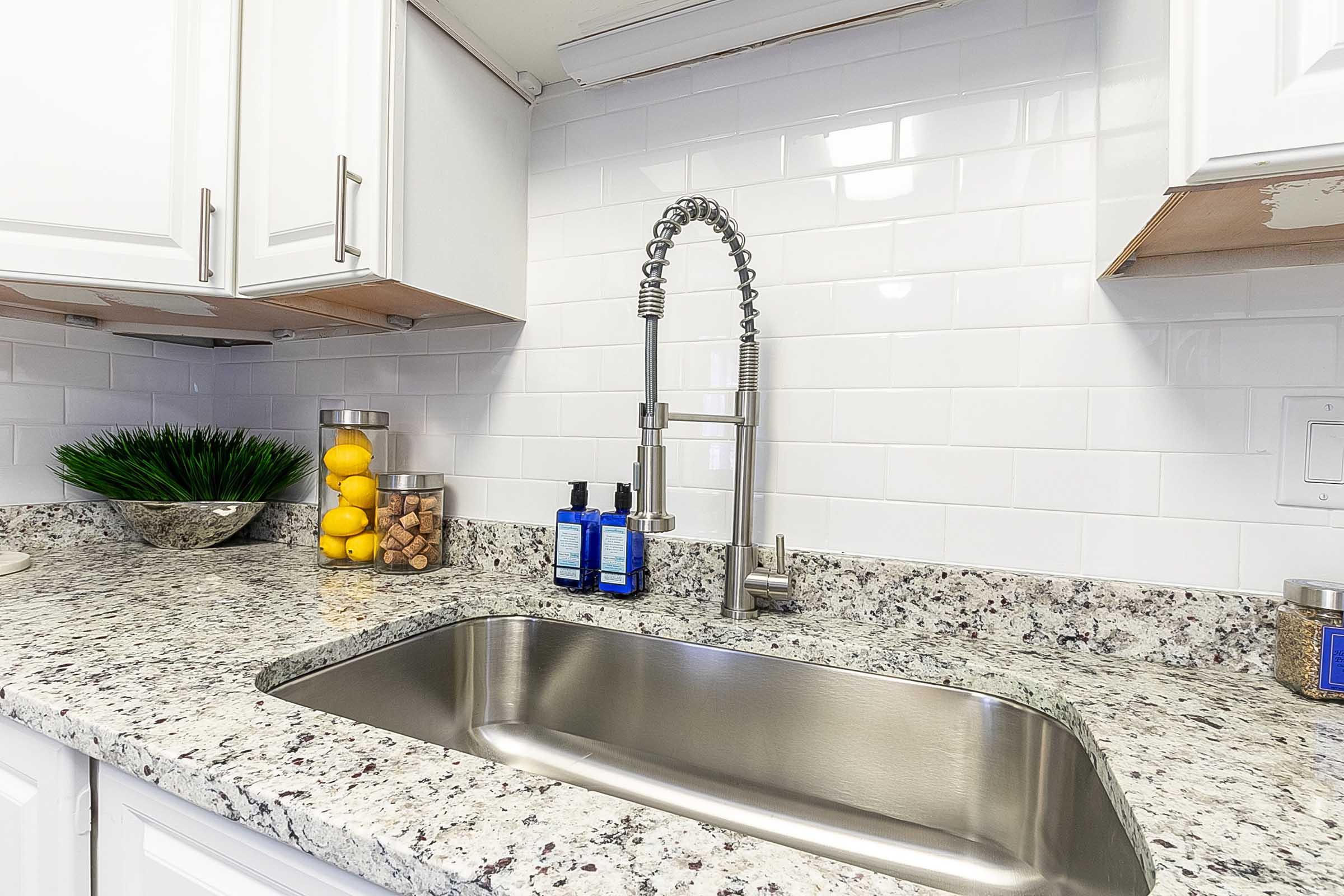 A modern kitchen sink with a stainless steel basin and a stylish pull-down faucet. The countertop is made of speckled granite, and there are decorative elements like a bowl of lemons and a glass jar filled with corks. White cabinets and a clean tiled backsplash complete the contemporary look.