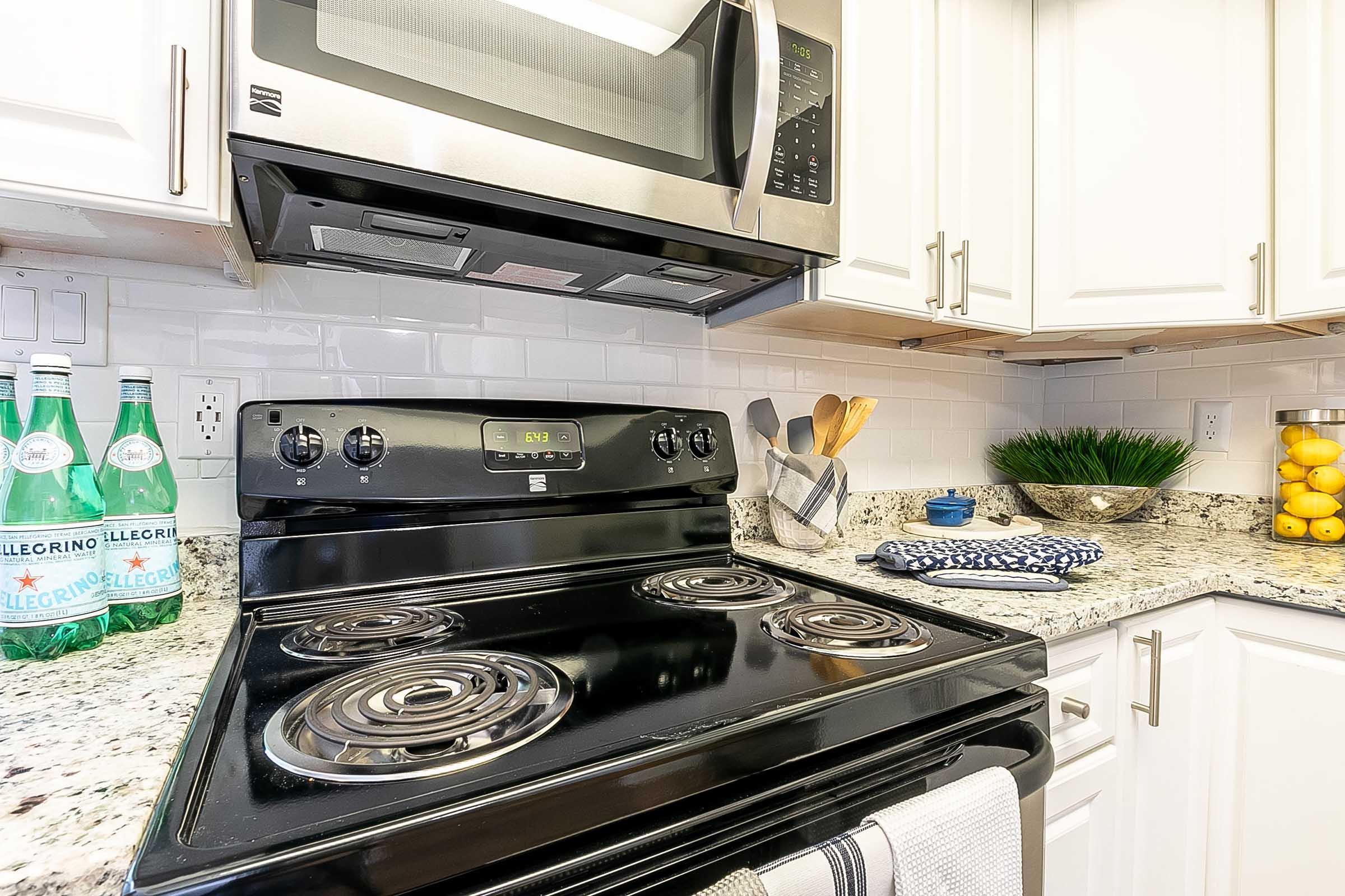 A modern kitchen featuring a black stovetop and built-in microwave. In the background, there are three bottles of sparkling water and a bowl of lemons on the counter. The countertops are made of granite, and there are utensils displayed in a container. The kitchen has white cabinetry and a bright ambiance.