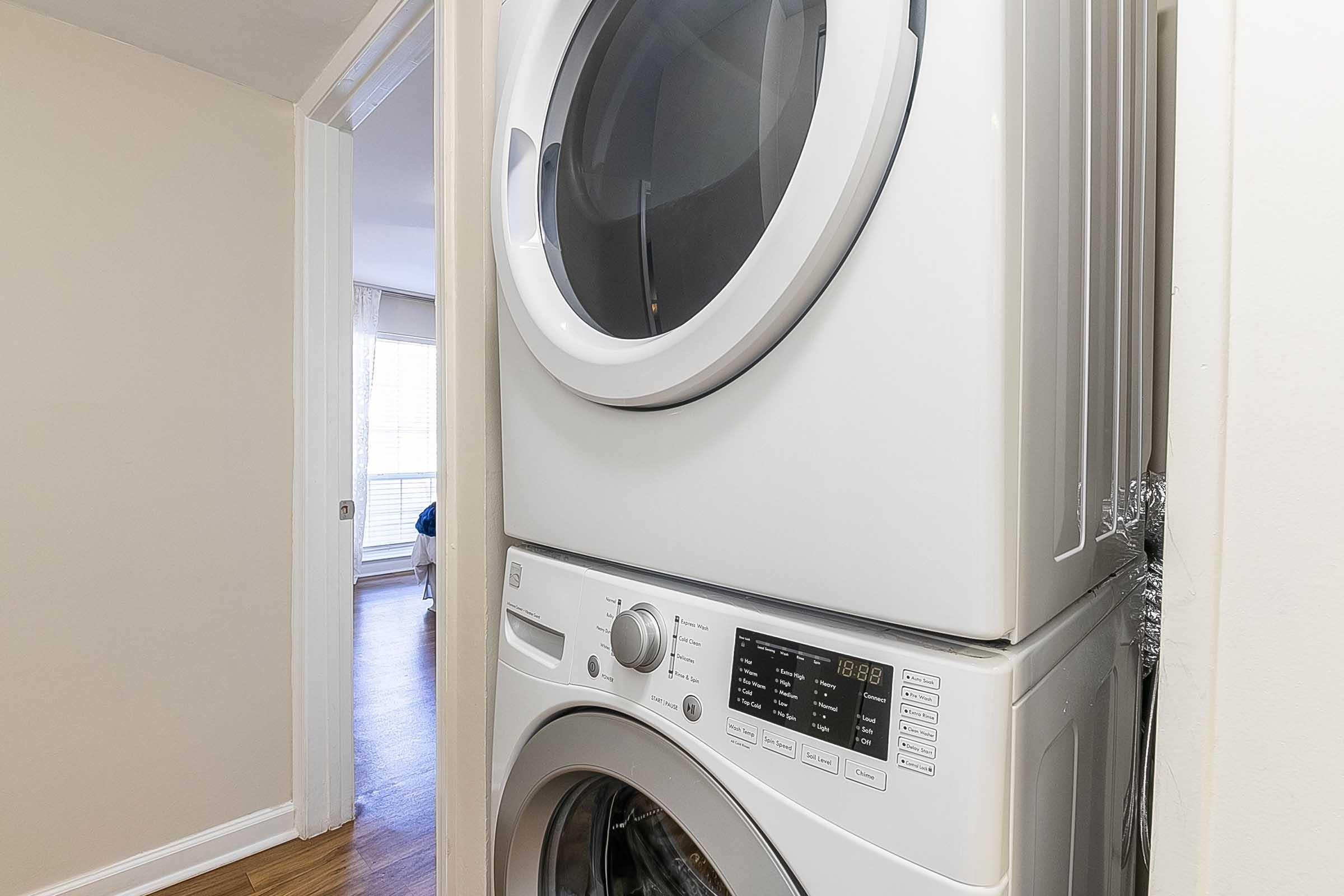 A compact laundry area featuring a stacked washer and dryer located in a narrow hallway. The walls are painted a light color, and there is a door leading to another room in the background with natural light coming through the window. The floor is hardwood, adding warmth to the space.