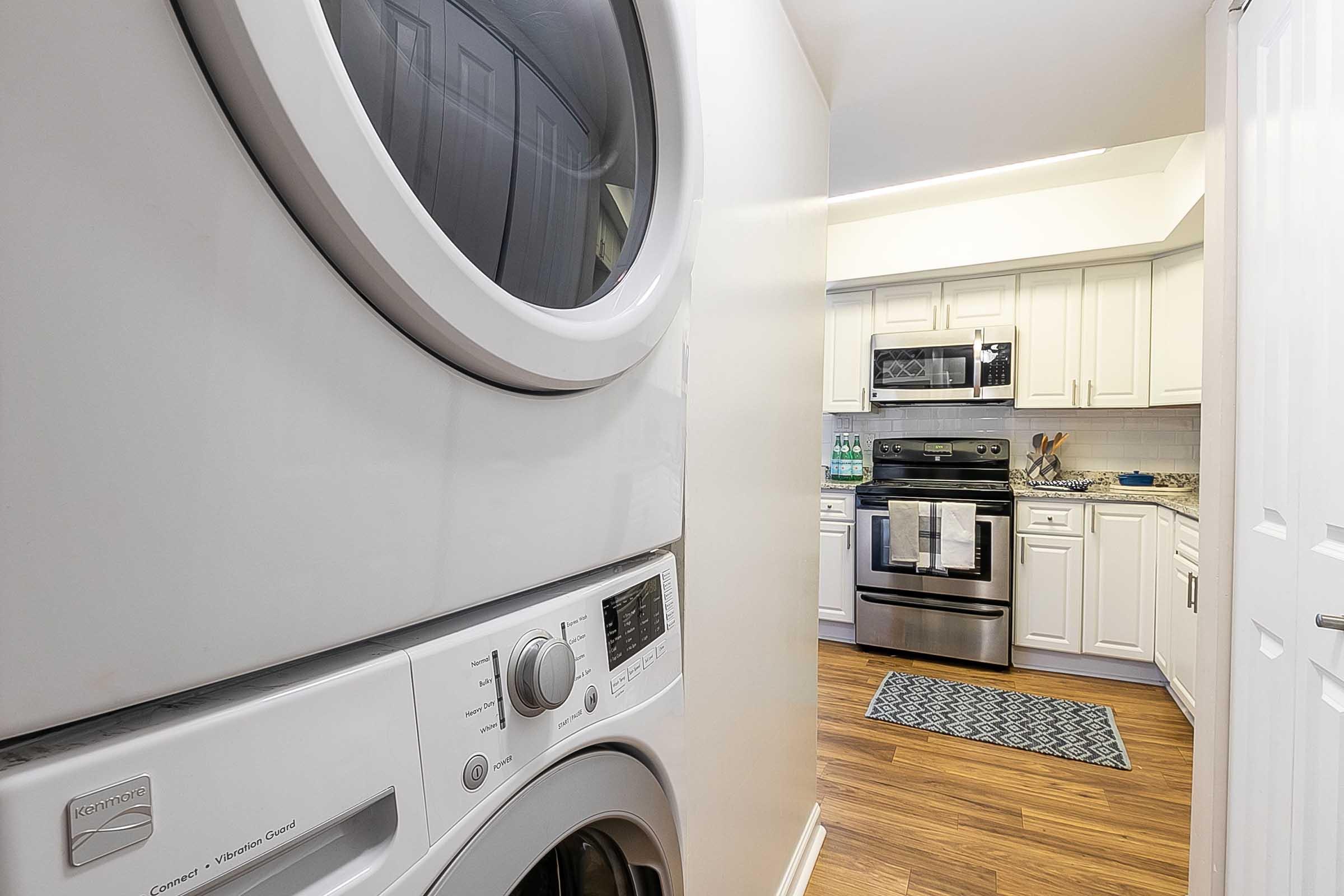 A laundry area featuring a white washing machine stacked with a dryer, viewed from a hallway. In the background, a modern kitchen can be seen with stainless steel appliances, white cabinets, and a blue patterned rug on the floor. The space is well-lit and tidy.
