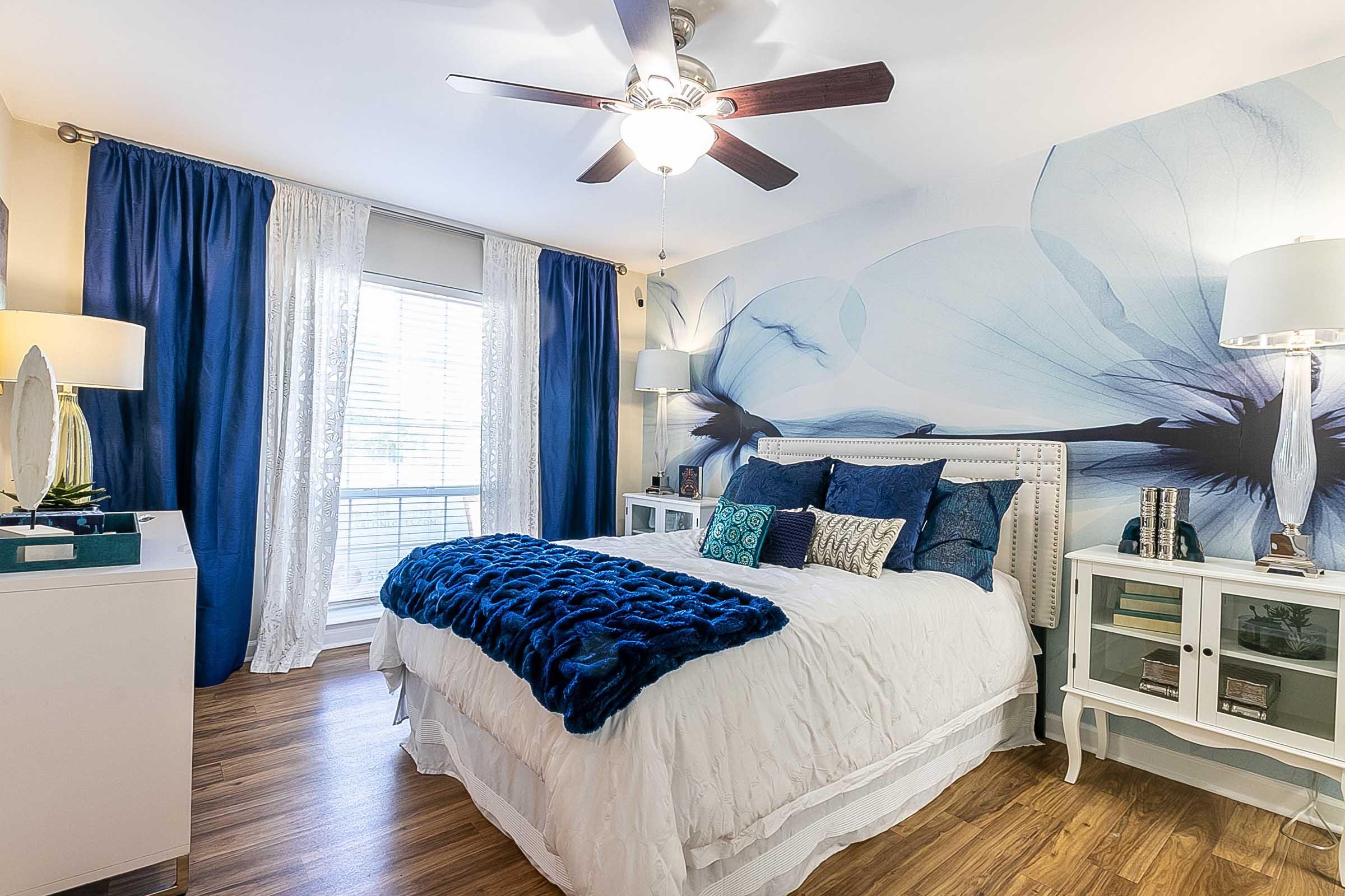 A cozy bedroom featuring a white bed covered with a blue throw blanket and an array of decorative pillows. The room has blue floral wall art, white curtains, and light wooden flooring. It includes two white nightstands with lamps, a dresser, and a window allowing natural light.
