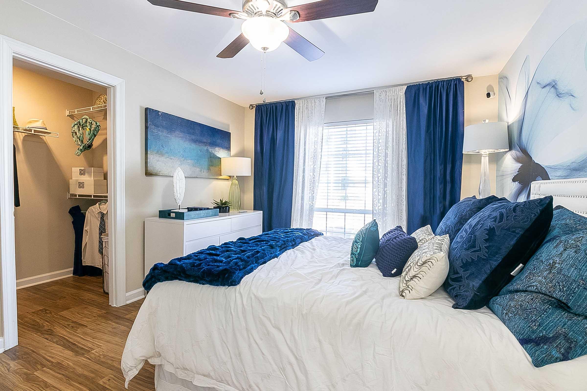 Cozy bedroom featuring a large bed with decorative pillows, a blue blanket, and stylish lamps. A ceiling fan hangs overhead. Light-colored walls complement navy curtains and artwork. A walk-in closet is visible through an open door, adding storage space and functionality to the room.
