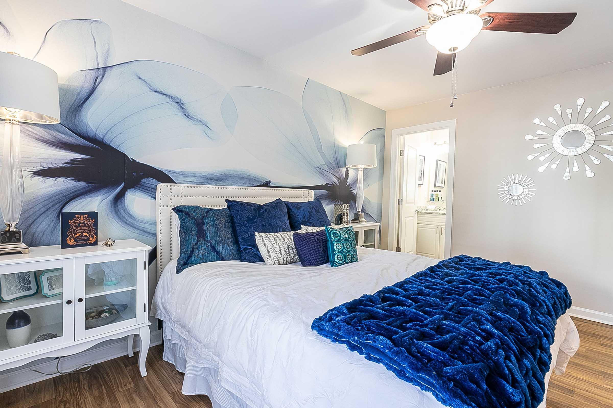 A cozy bedroom featuring a white bed with fluffy blue pillows and a textured blue blanket. The walls are adorned with a floral mural in shades of blue, and there are stylish nightstands with lamps on either side. A bright bathroom is visible in the background, adding to the serene atmosphere.