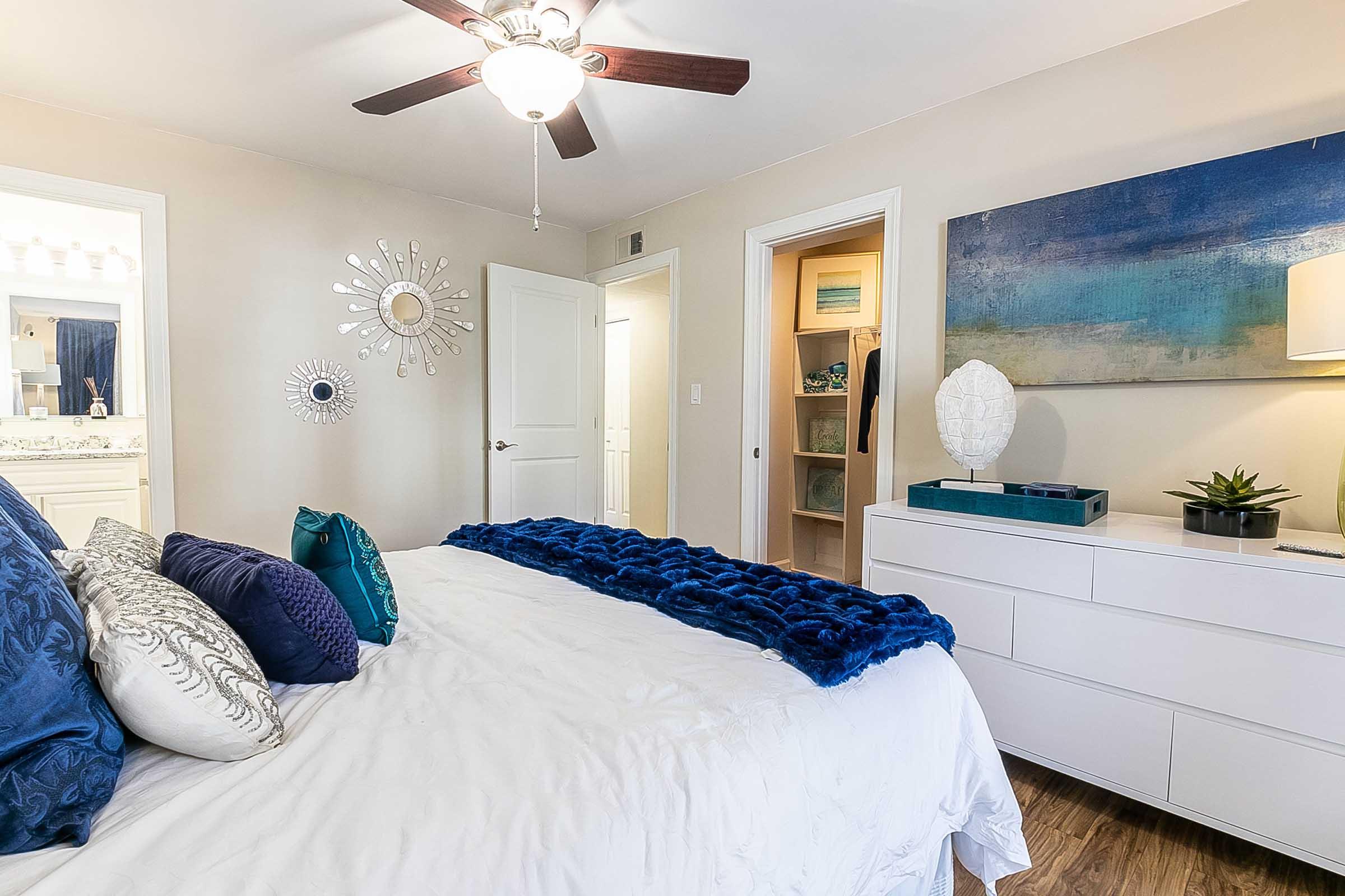 A cozy bedroom featuring a white bed with decorative blue pillows and a plush blue throw. There's a ceiling fan, a modern dresser, and artwork on the wall. A door leads to a bathroom, and another door opens to a closet. The overall decor is inviting and stylish, with a soothing color scheme.