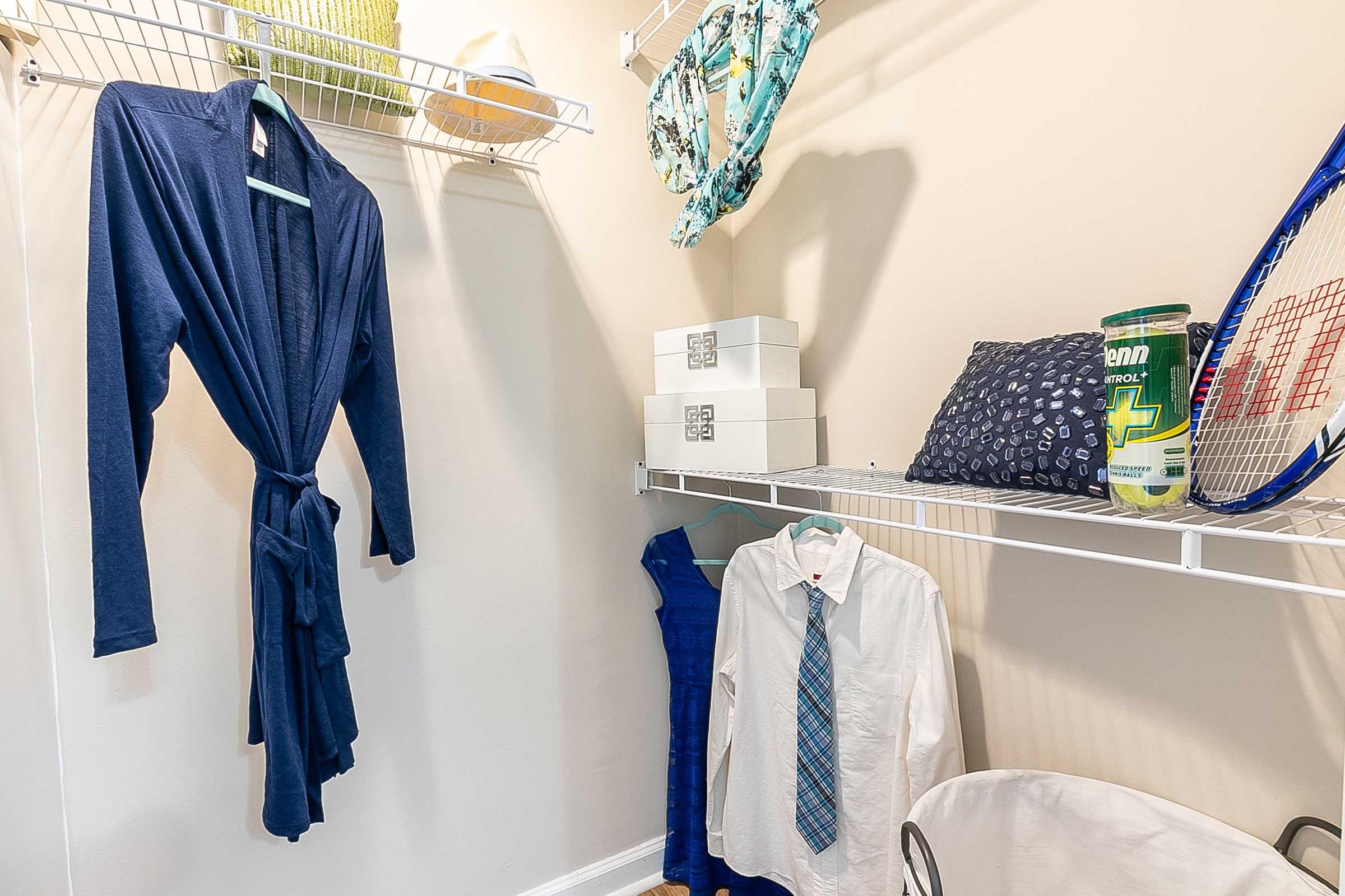 A tidy closet space featuring a navy blue bathrobe on a hanger, a white dress shirt with a tie, storage boxes, and a canister on a shelf. A tennis racket is also visible, along with some folded items and a green object in the corner. Light-colored walls enhance the organized atmosphere.