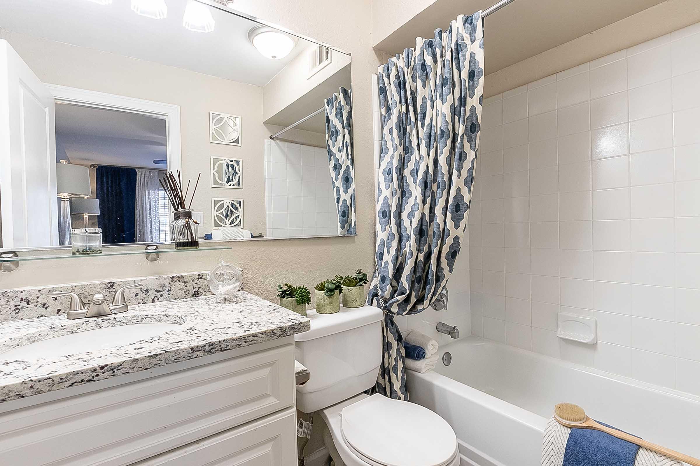 A modern bathroom featuring a bathtub, white cabinets, and granite countertop. A colorful shower curtain with blue flowers adds a decorative touch, while potted plants and toiletries are neatly arranged. A mirror reflects the space, enhancing the room's brightness. Soft lighting complements the neutral color scheme.