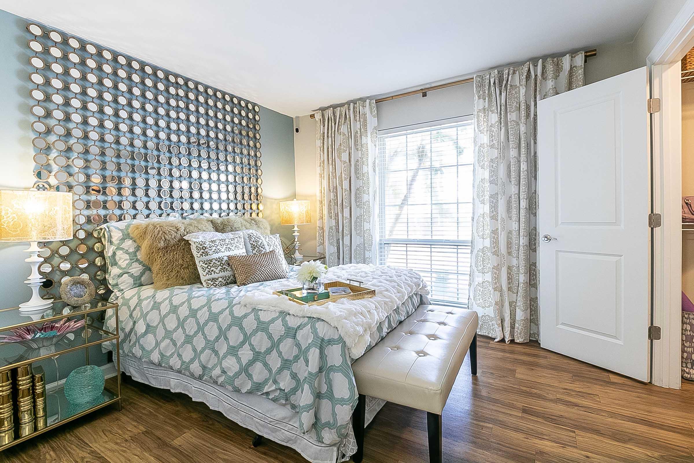 Cozy bedroom with a decorative wall featuring circular mirrors, a queen-sized bed with plush bedding, and a beige upholstered bench at the foot. There are bedside tables with lamps, light curtains framing a window, and warm wood flooring, creating a welcoming and stylish atmosphere.