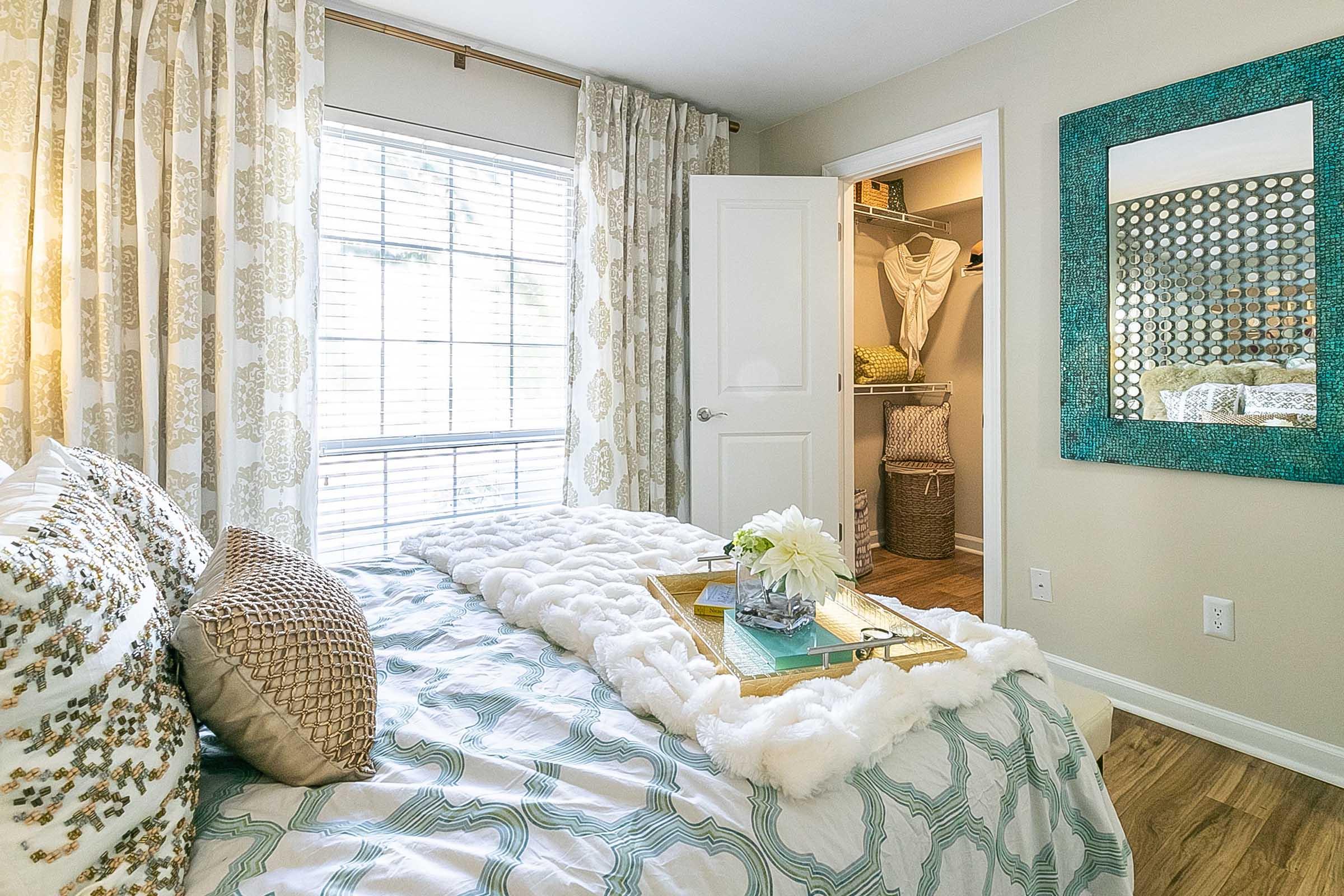 A cozy bedroom featuring a bed with a fluffy white throw, decorative pillows, and a tray with books and flowers. Natural light streams through a window adorned with patterned curtains. A mirror hangs on the wall, and an open closet reveals woven storage baskets.