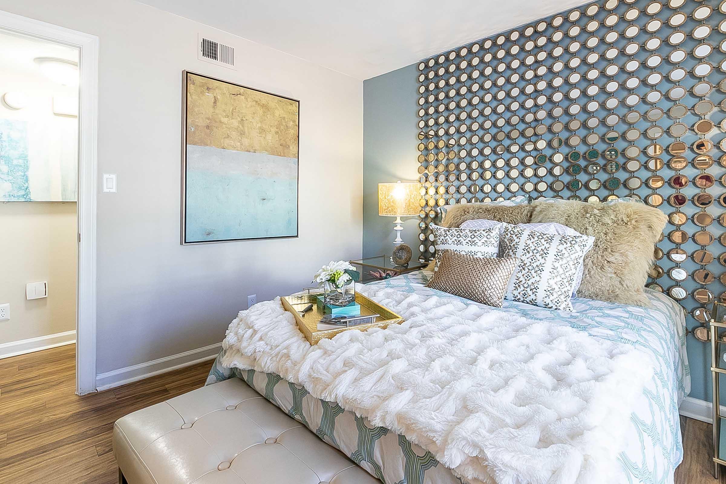 A cozy bedroom featuring a bed with a plush white blanket and decorative pillows. A wall adorned with circular mirrors creates a unique accent. A bedside table with a stylish lamp and a tray holding flowers sits beside the bed. The room has warm wood flooring and a calming color scheme of blues and neutrals.