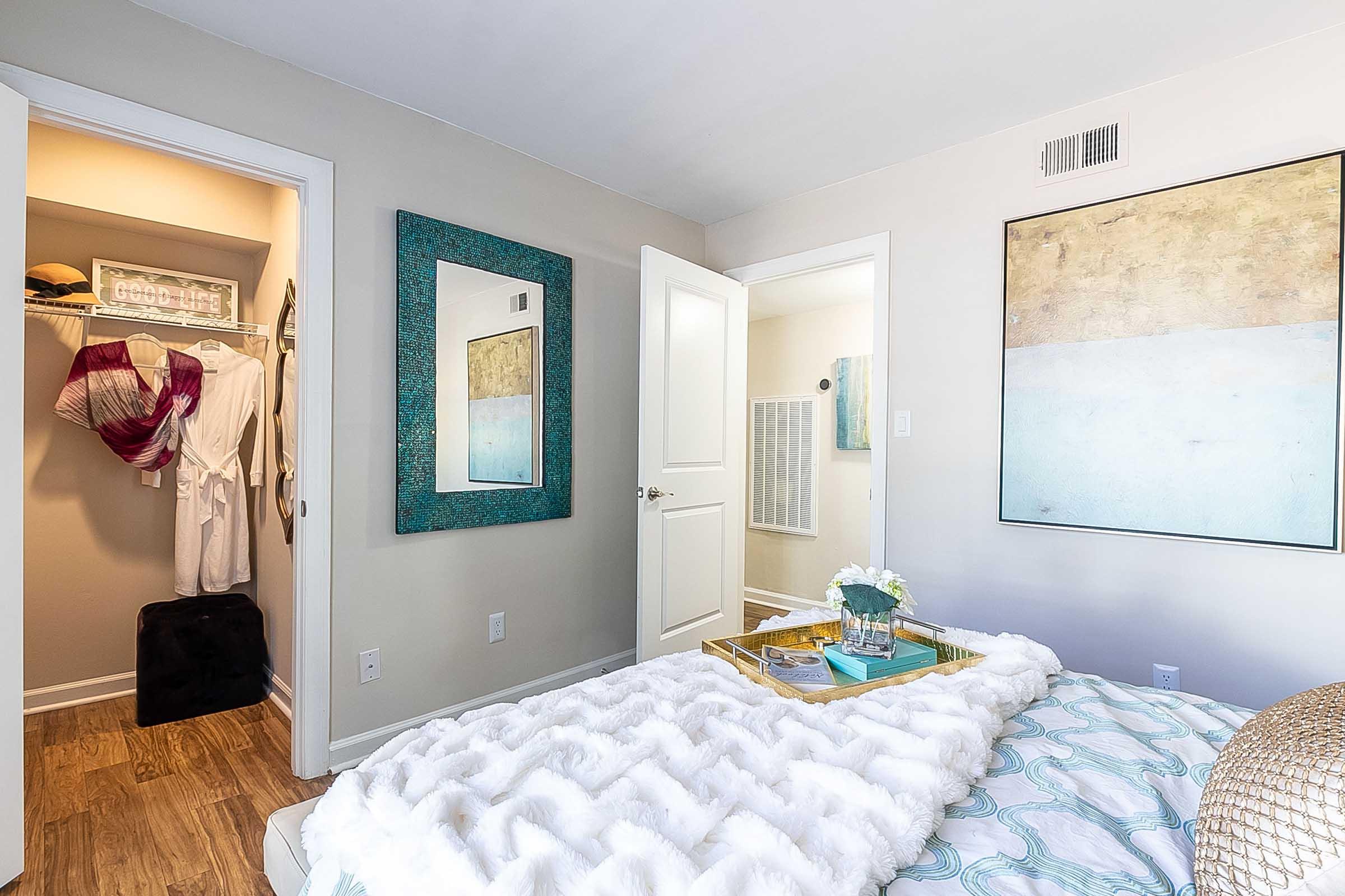 A cozy bedroom with a neatly made bed covered in a textured white blanket. A decorative tray with a vase of flowers sits on the bed. A large mirror reflects the room, and there is an open closet showing neatly hung clothes. A doorway leads to a bathroom in the background, enhancing the room's inviting ambiance.