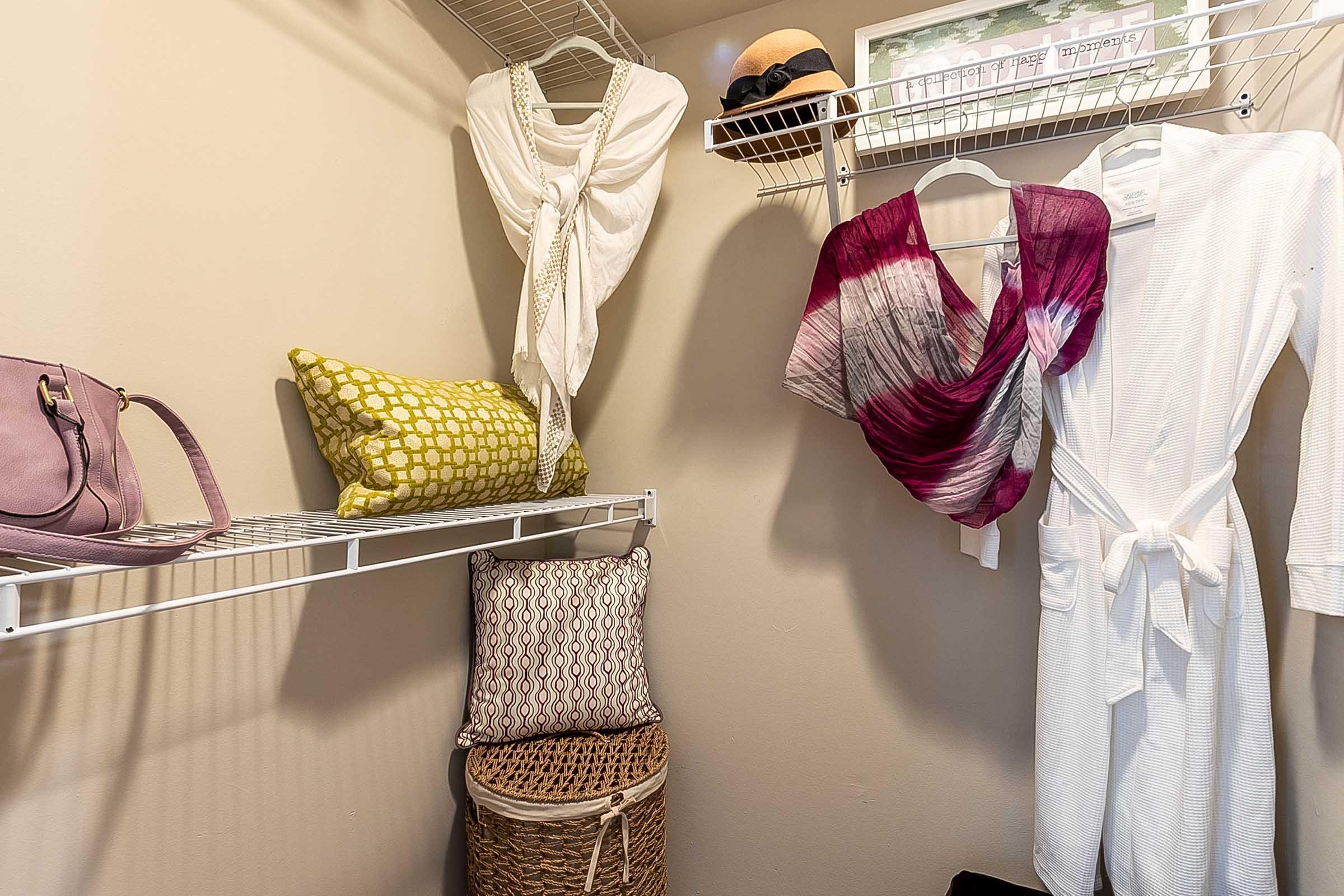 A neatly organized closet featuring two hanging scarves, a white robe, and several decorative pillows on shelves. There's a woven basket on the floor and a handbag hanging from a shelf, all set against a neutral wall. The space is bright and inviting, showcasing a minimalist style.