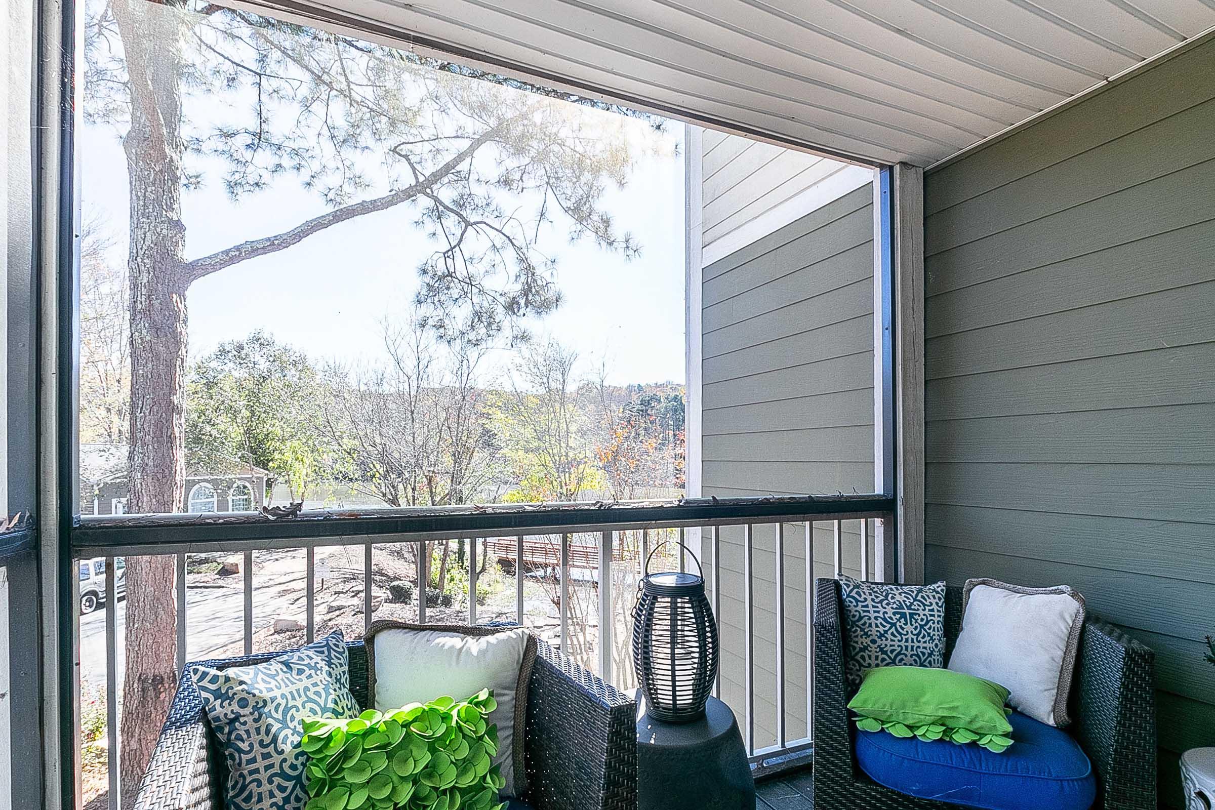 A cozy balcony view featuring two wicker chairs adorned with colorful cushions, a decorative lantern, and a glimpse of trees and greenery outside. The space is brightened by natural light, creating a welcoming atmosphere for relaxation and enjoyment of the outdoors.