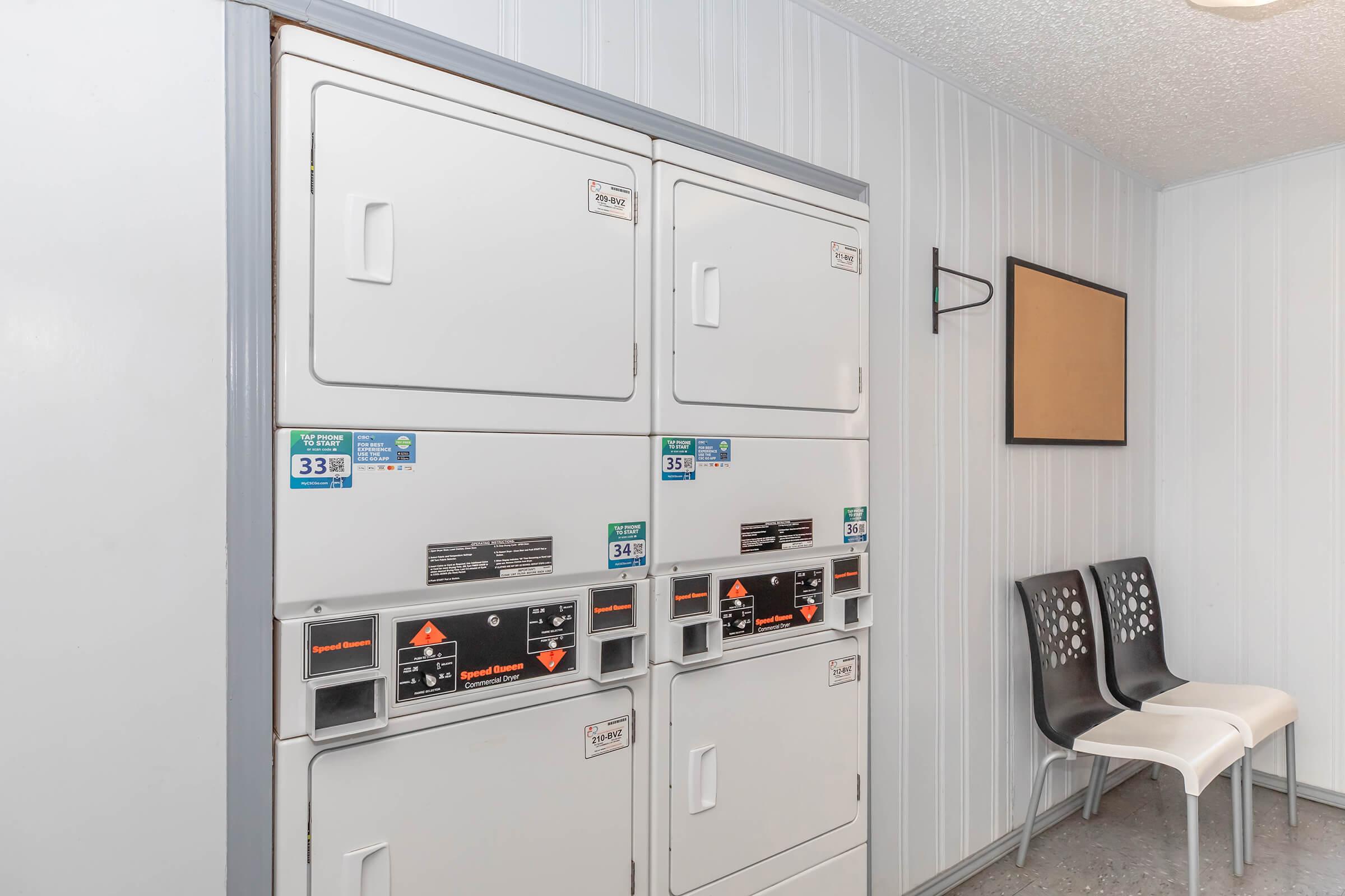 A row of stacked coin-operated laundry machines in a clean, bright laundry room. To the right, there are two modern chairs and a bulletin board mounted on the wall, all set against a light-colored paneling backdrop.