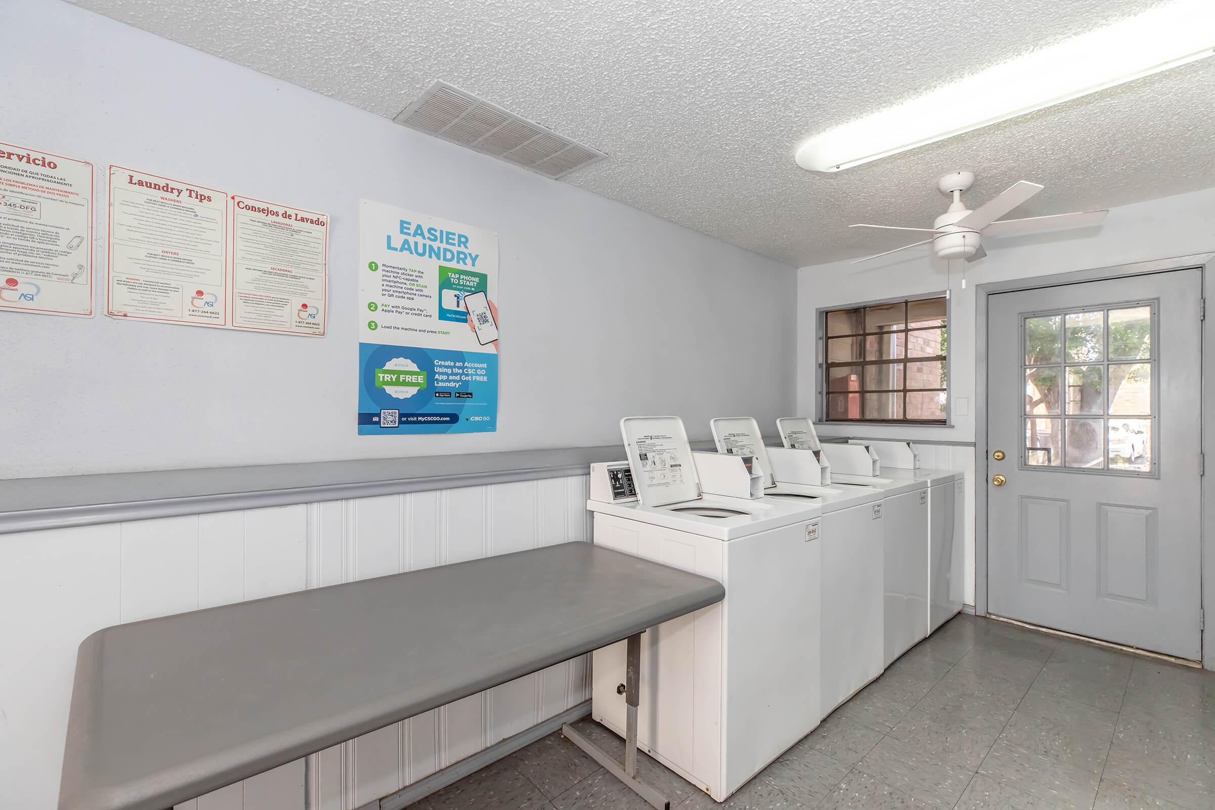 A small laundry room with a folding table, several washing machines, and a door leading outside. The walls are painted in light colors, and posters are displayed with laundry tips. A ceiling fan is visible, along with a window that provides natural light.