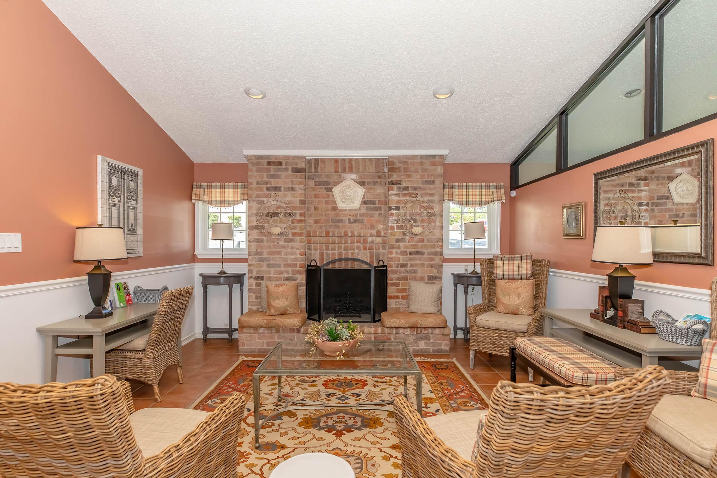 A cozy living room featuring a brick fireplace at the center. The walls are painted in a warm peach tone, and there are two comfortable wicker chairs with patterned cushions. A glass coffee table sits on a decorative rug, and small side tables flank the fireplace. Large windows allow natural light to brighten the space.