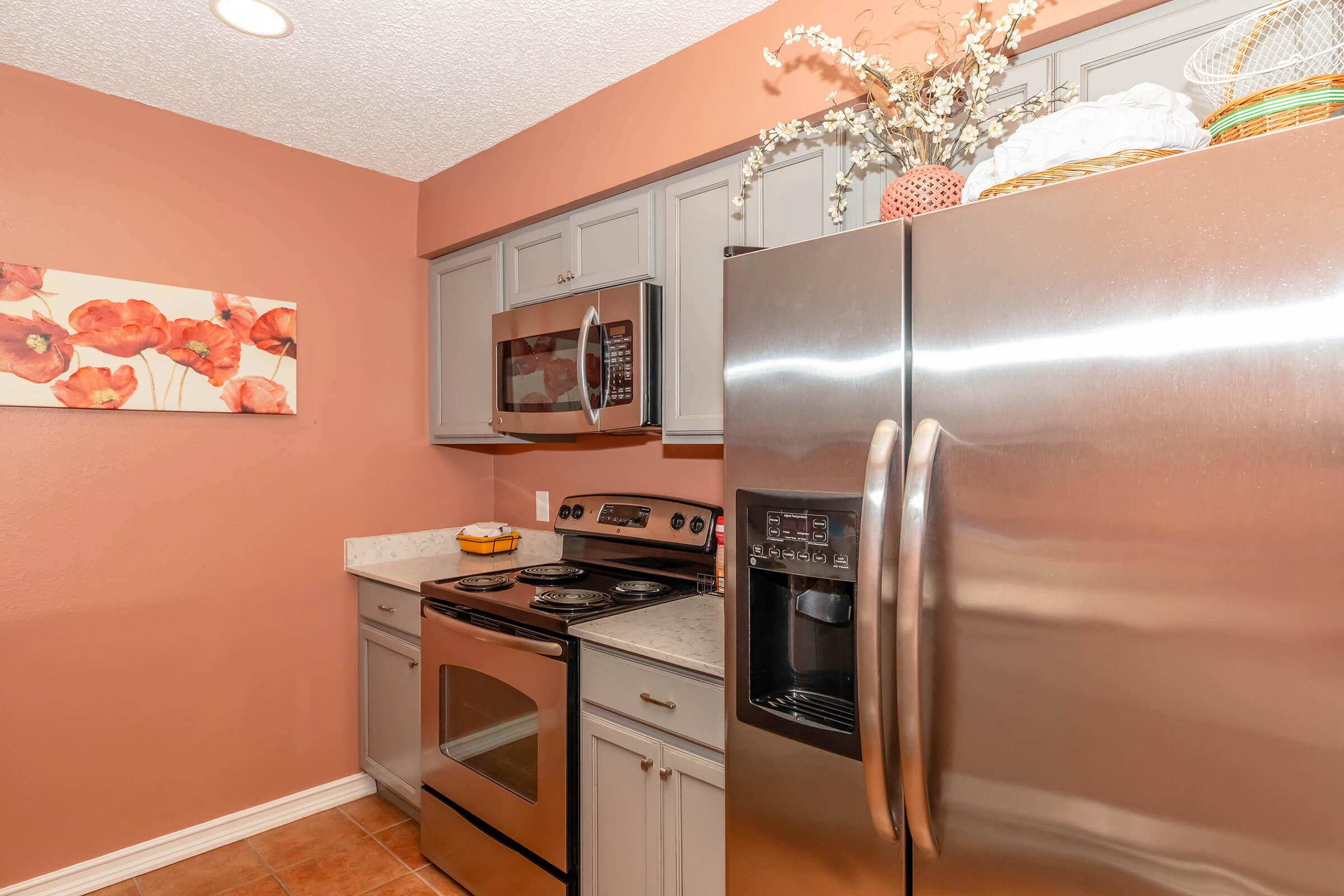 Modern kitchen featuring stainless steel appliances including a refrigerator, microwave, and stove. The cabinets are gray with a warm orange wall adorned with a floral artwork. Countertops have a light natural stone appearance, creating a cozy and stylish atmosphere.