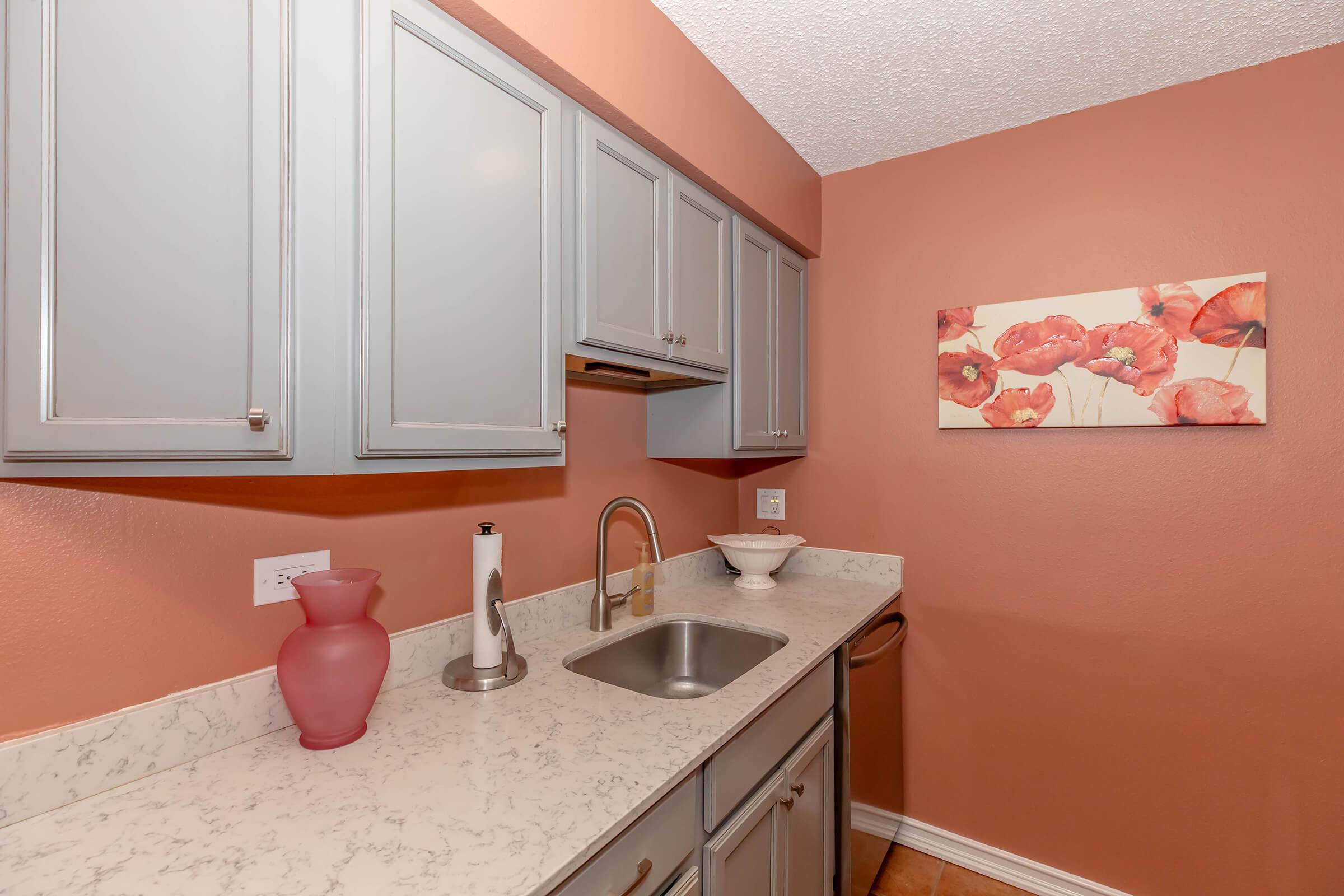 A modern kitchen space featuring light gray cabinets, a marble countertop, and a stainless steel sink. The walls are painted a warm peach color, complemented by a pink vase on the countertop and a floral artwork depicting red poppies.