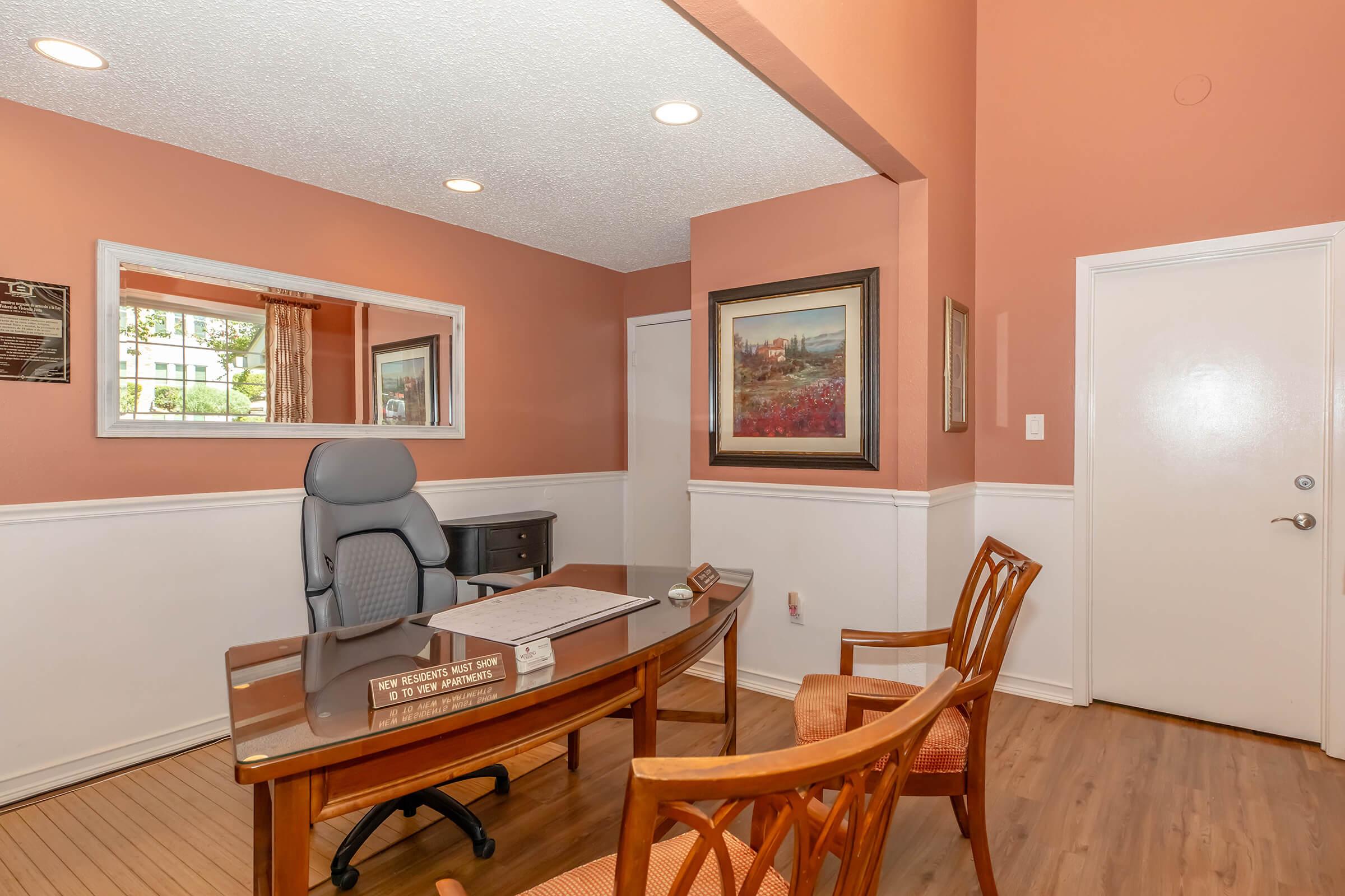 Interior of an office space featuring a wooden desk with a nameplate, an ergonomic office chair, and two wooden chairs. The walls are painted in a warm peach color, complemented by white trim. A mirror and a framed painting are visible on the walls, along with natural light coming from a window.