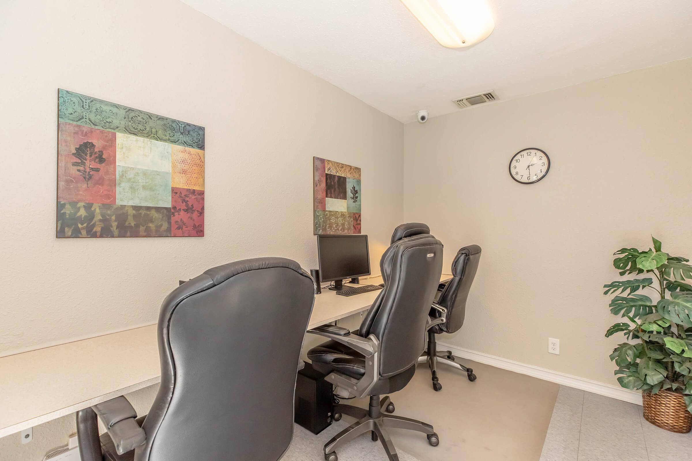 A small office space with four ergonomic black chairs around a light-colored desk. Two decorative wall art pieces hang on the beige walls, and a clock is mounted on the right side. A green potted plant adds a touch of greenery to the room, which has a neutral and professional atmosphere.