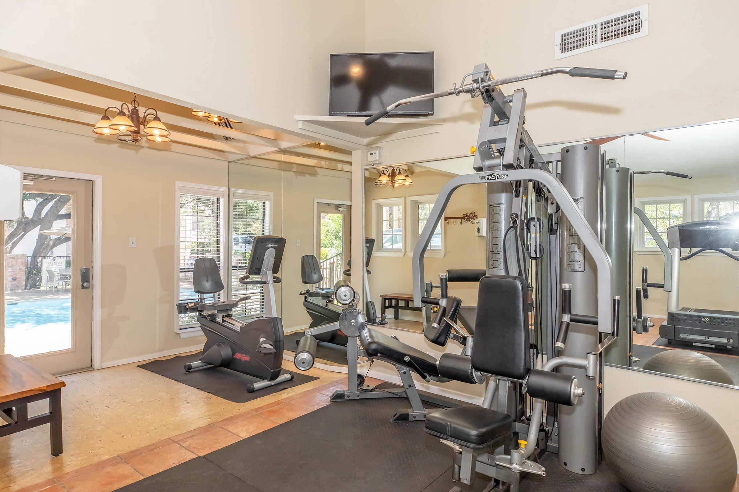 A home gym featuring various fitness equipment including a weight machine, exercise bike, and a stability ball. There is a large mirror on one wall and a TV mounted above. Natural light comes in through sliding glass doors leading to a pool area. The floor is a mix of tile and rubber matting.