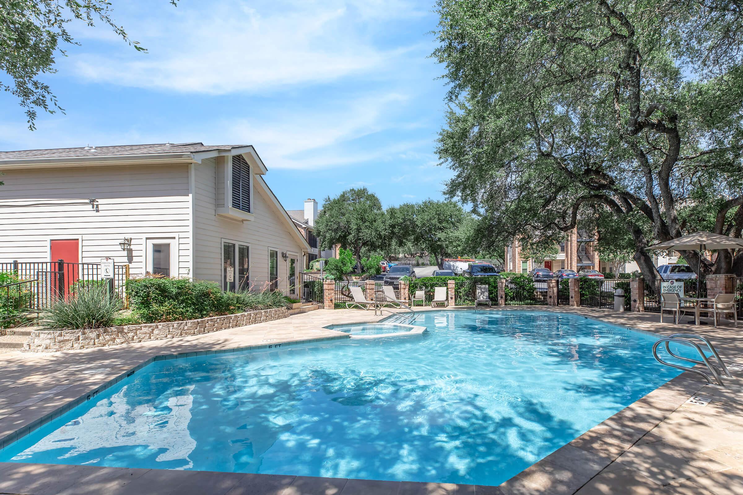 A serene outdoor swimming pool surrounded by lush greenery and a patio area. The pool features a clear blue water surface, with lounge chairs and an umbrella nearby. In the background, a residential building and various trees are visible under a clear blue sky, creating a relaxing atmosphere.