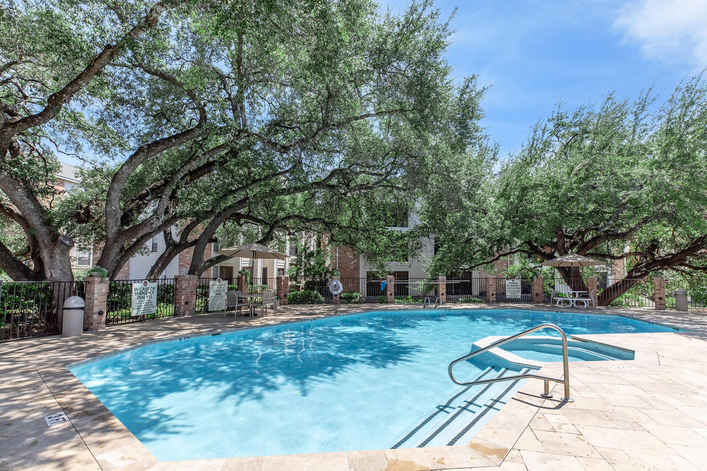 A serene swimming pool surrounded by lush, green trees. The pool has a gentle curve and a staircase leading in. There are lounge chairs and umbrellas nearby, with a clear blue sky overhead, creating a relaxing outdoor atmosphere.