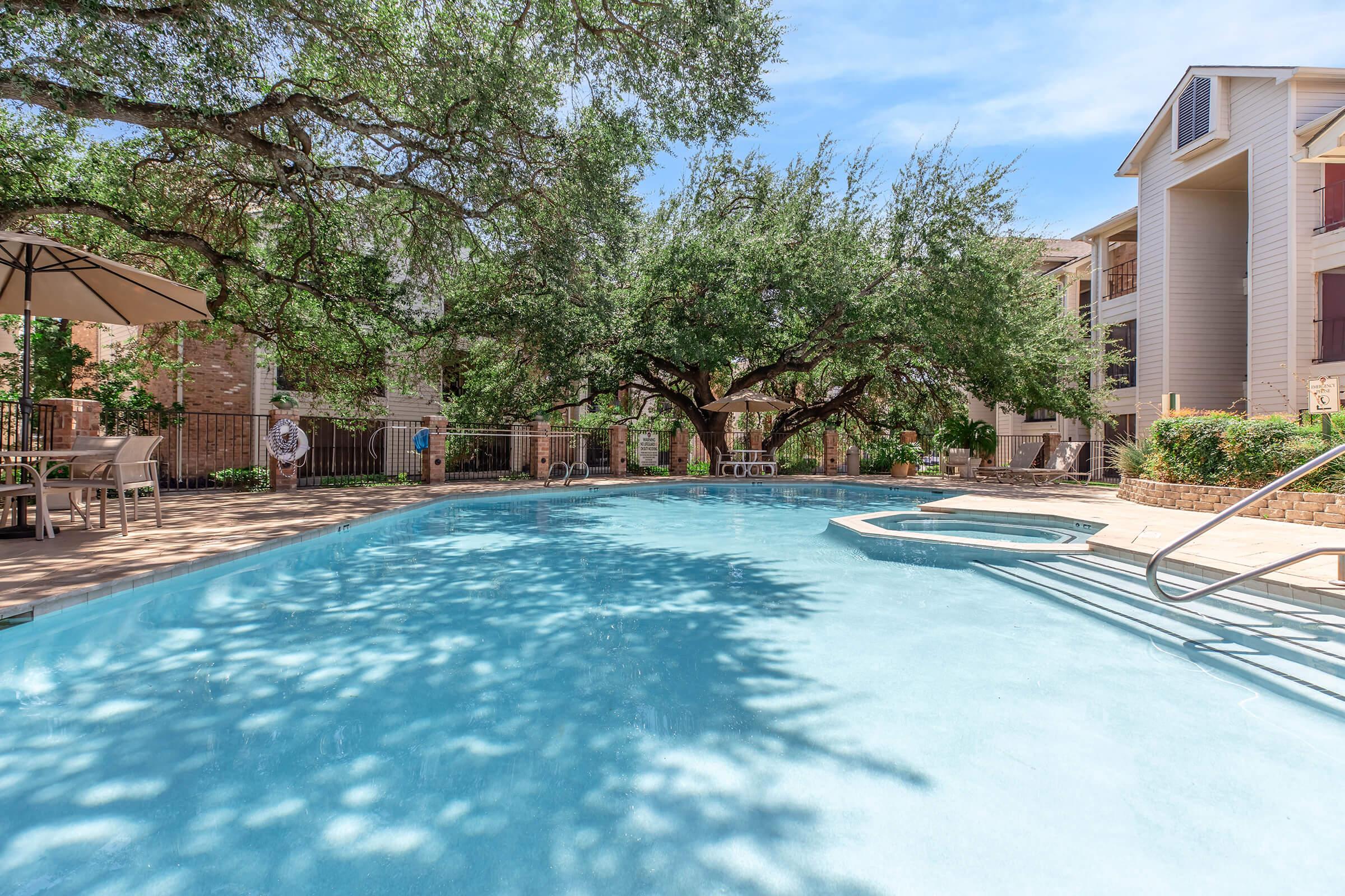 A serene swimming pool surrounded by lush greenery and shade from large trees. Sunlight filters through the leaves, casting playful shadows on the water. The area features a spacious patio with lounge chairs and a shaded umbrella, creating a relaxing outdoor environment.