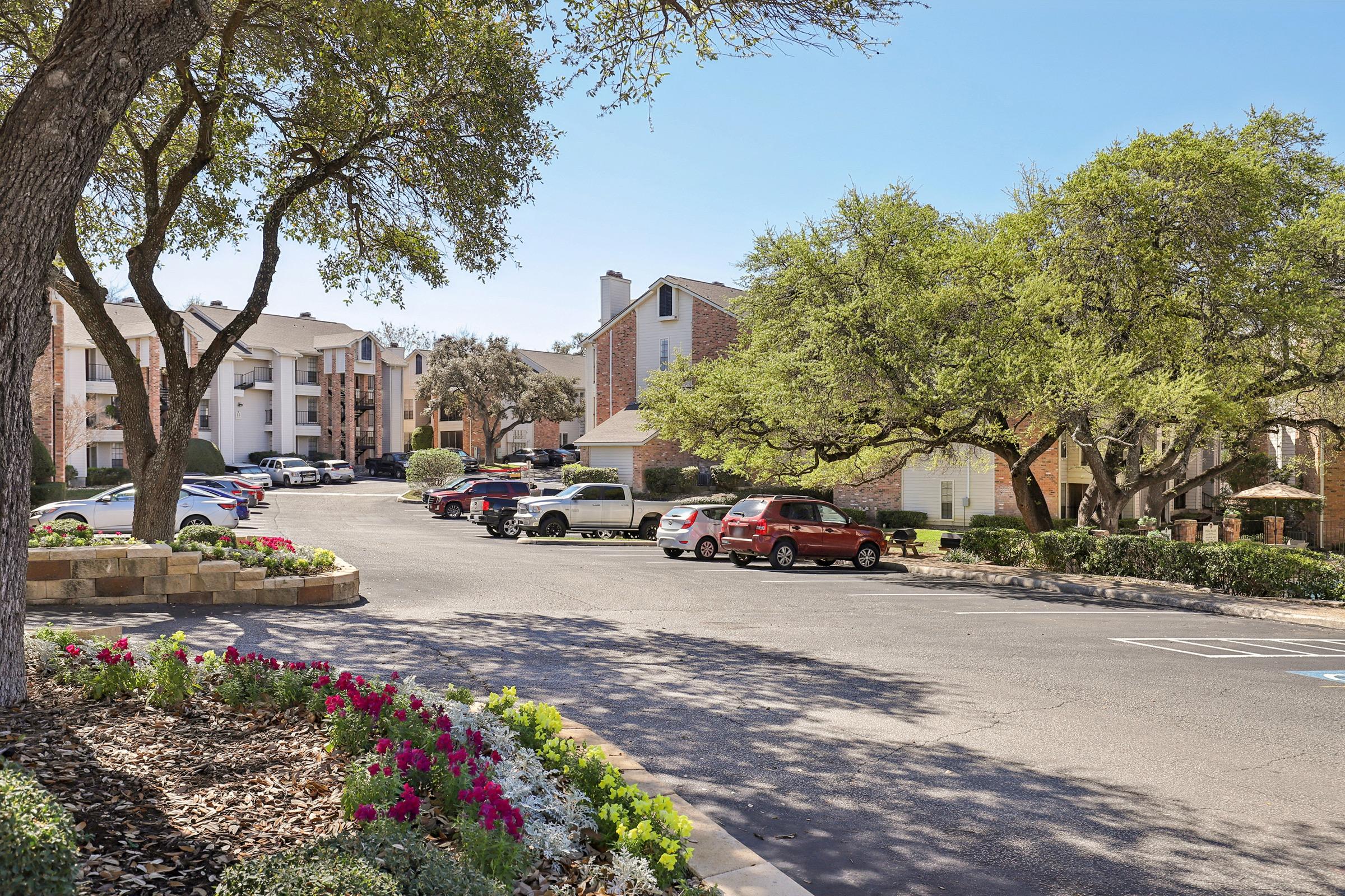 A residential area featuring multiple apartment buildings surrounded by green trees. The scene includes a paved parking lot with several vehicles and flower beds with colorful blooms in the foreground, all under clear blue skies.