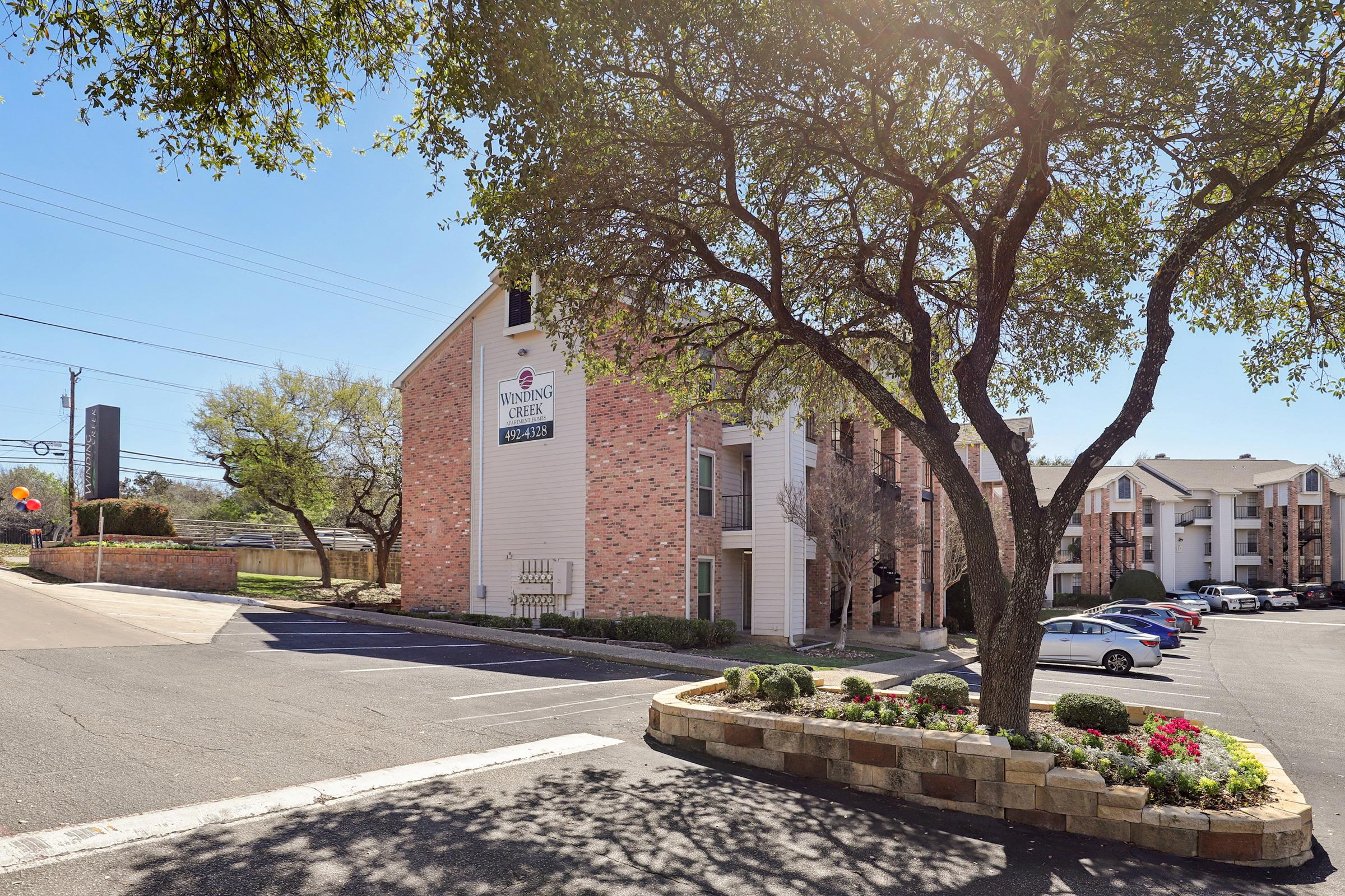 A view of the exterior of a lodging facility, featuring a brick building with a sign that reads "Wyndham Gardens." In the foreground, there is a landscaped area with flowers and shrubs, along with several parked cars in the lot. The scene is bright and sunny, with trees providing shade.