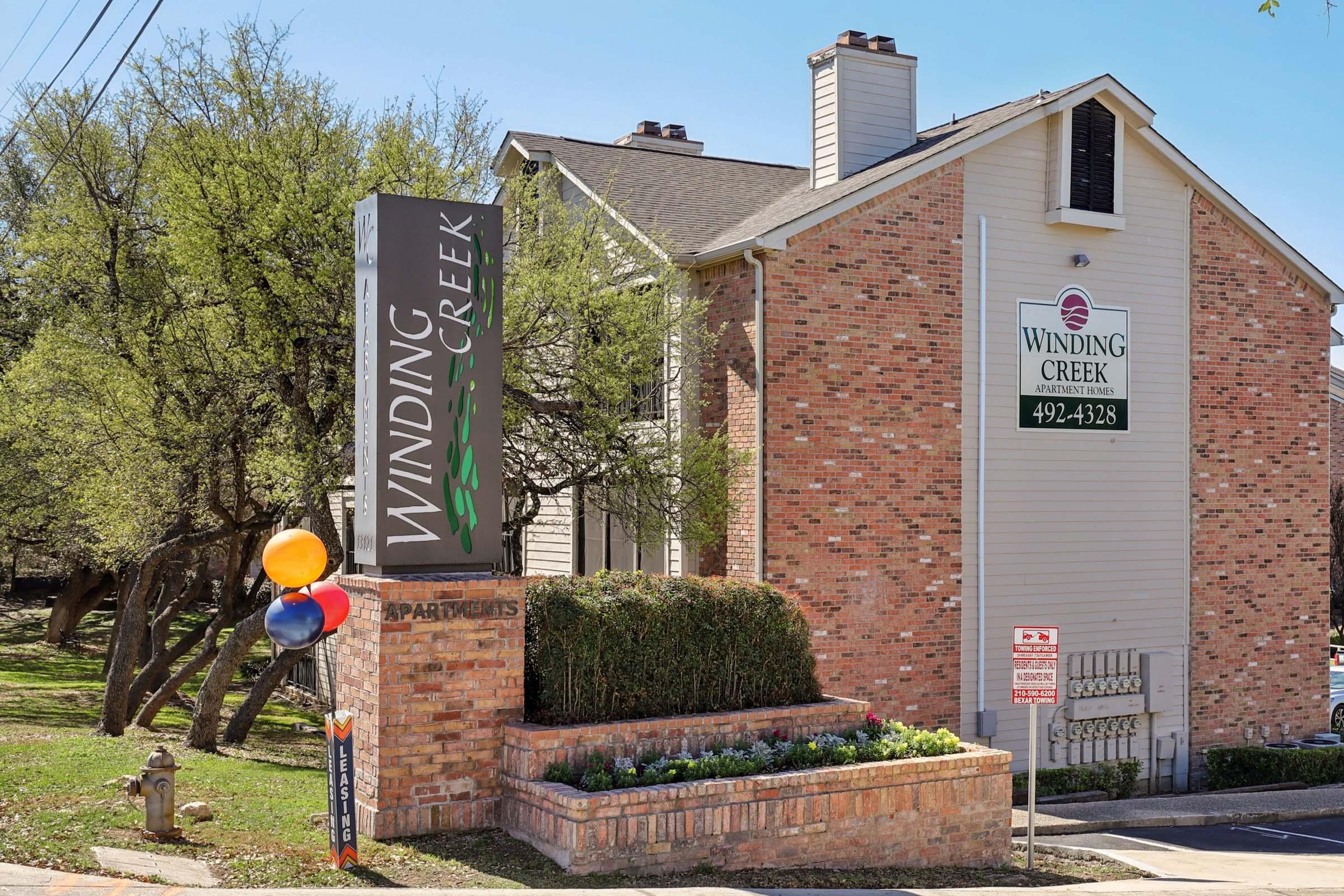 Sign for Winding Creek Apartments in a residential area, featuring a modern design with vibrant colors. The building is brick, surrounded by trees and landscaping. A cluster of colorful balloons is in front of the sign, adding a festive touch.