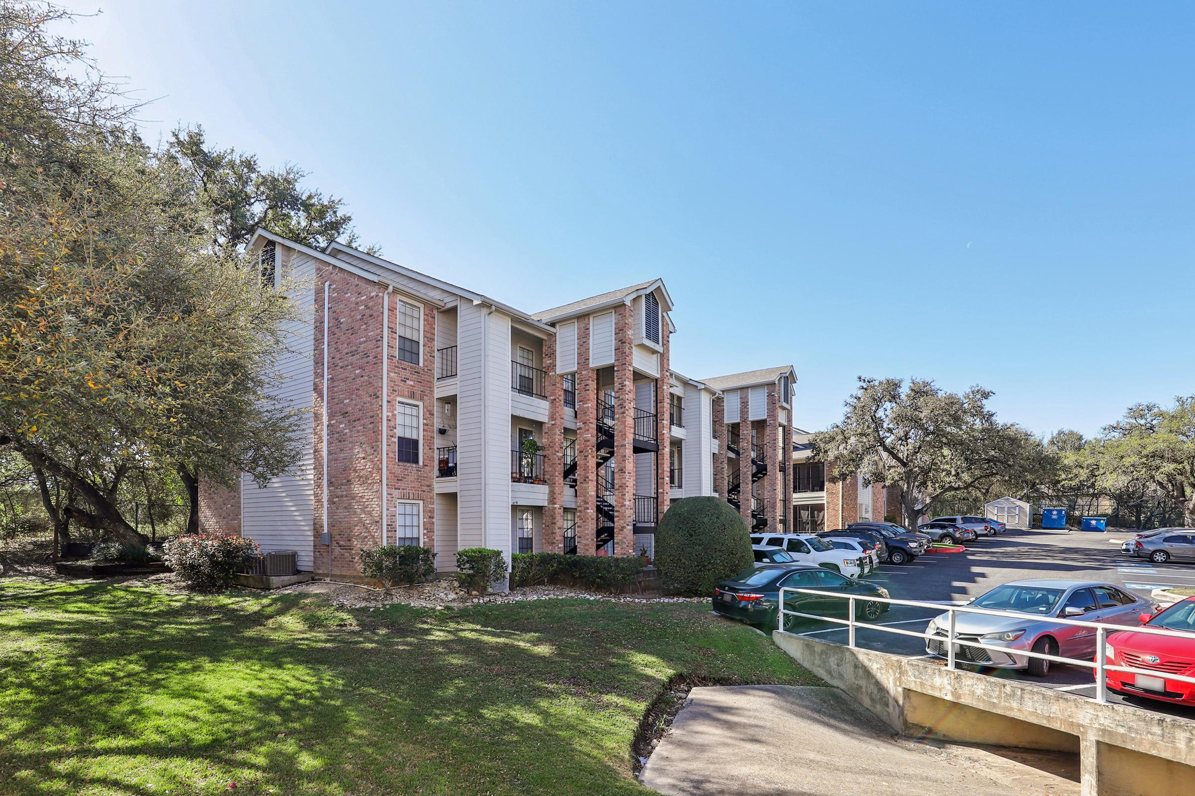 A multi-story apartment building with a brick facade and white siding, surrounded by greenery. The parking lot in front has several cars parked. Clear blue sky above, with a few trees visible in the area.