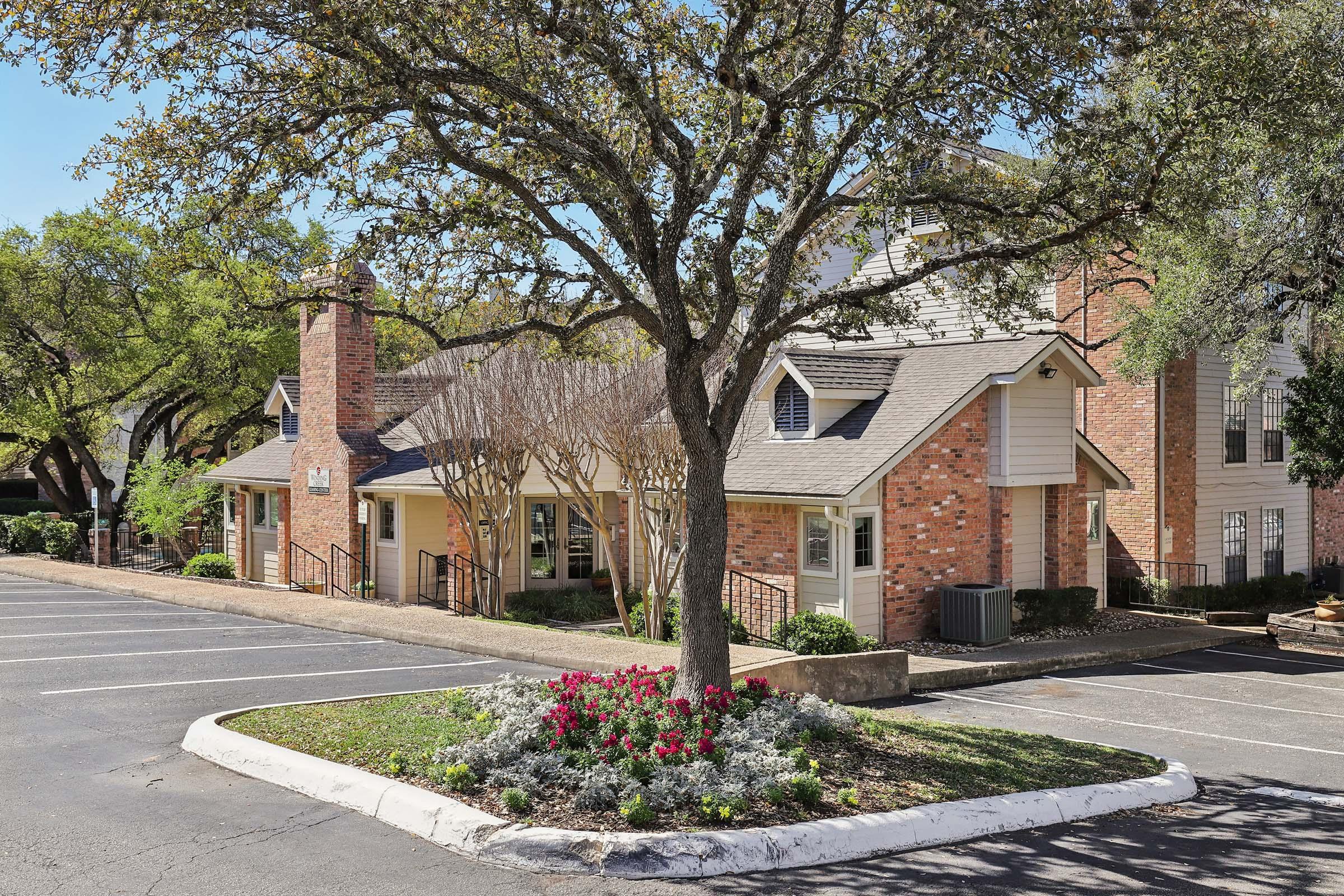A landscaped area showing a brick building with a sloped roof, surrounded by trees. In the foreground, there is a circular flower bed with blooming flowers. The parking lot is empty, and blue sky is visible above the scene.