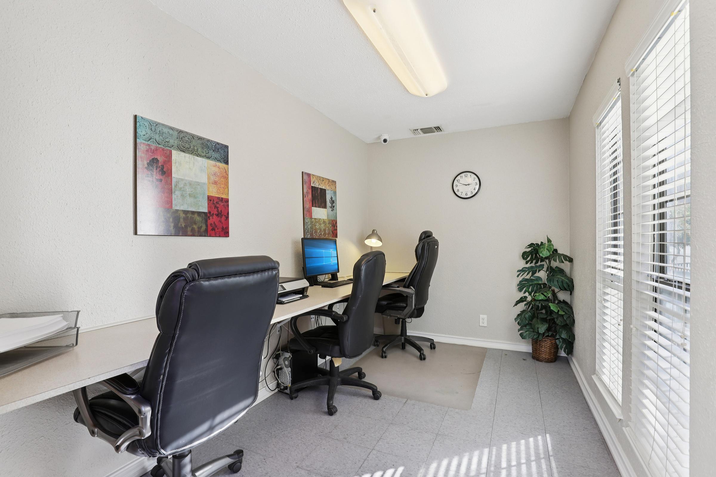 A bright and spacious office space featuring three black office chairs at white desks with computers. There are colorful wall art pieces on one wall, a clock on another, and a potted plant in the corner. Large windows provide natural light, enhancing the workspace ambiance.