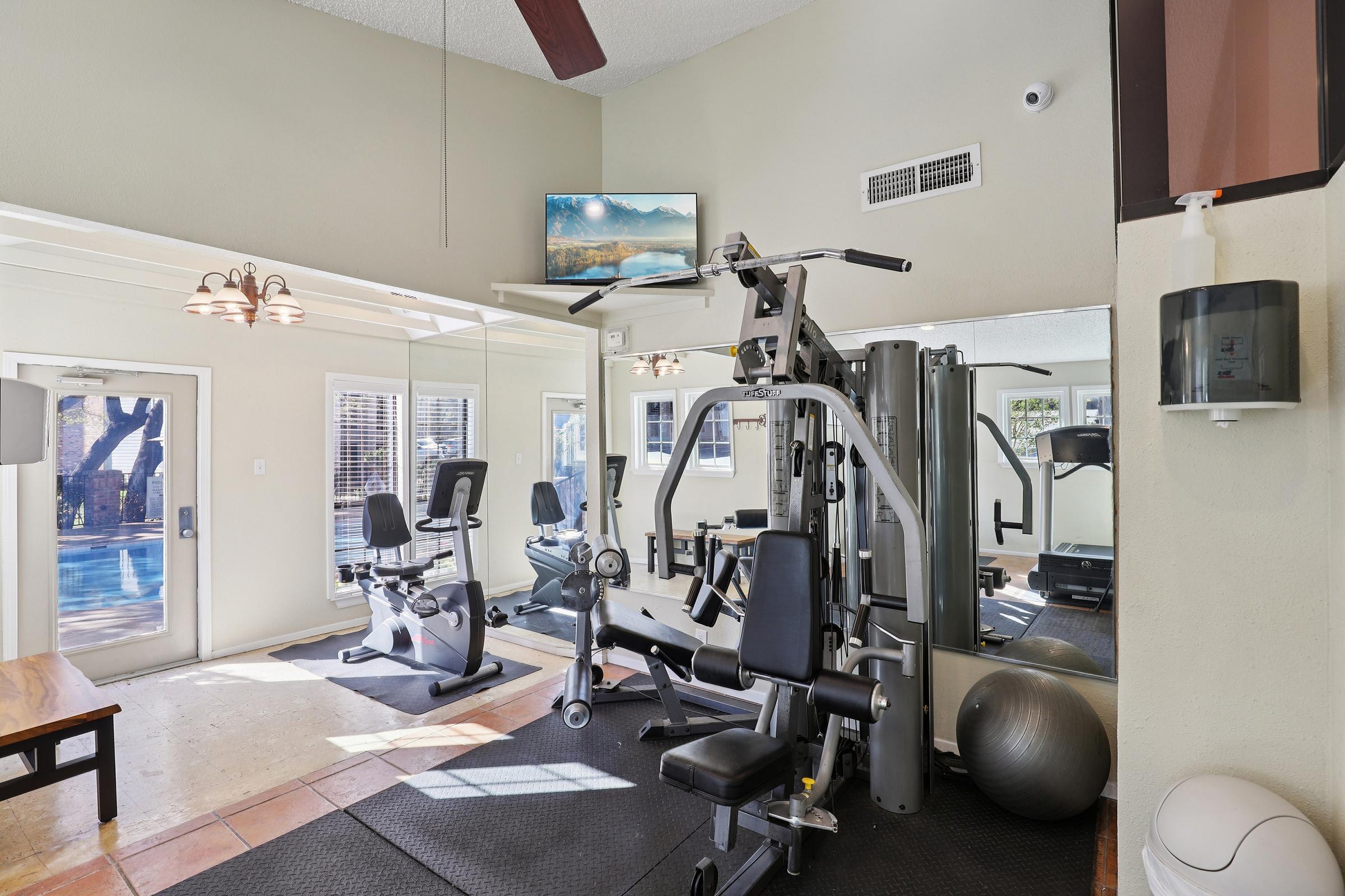 A well-equipped home gym featuring a multi-station weight machine, exercise bike, and a stability ball. There's a large mirror on one wall reflecting the equipment, and a flat-screen TV mounted above. Natural light comes from large windows overlooking a pool area. The space has a bright, airy feel with neutral colors.
