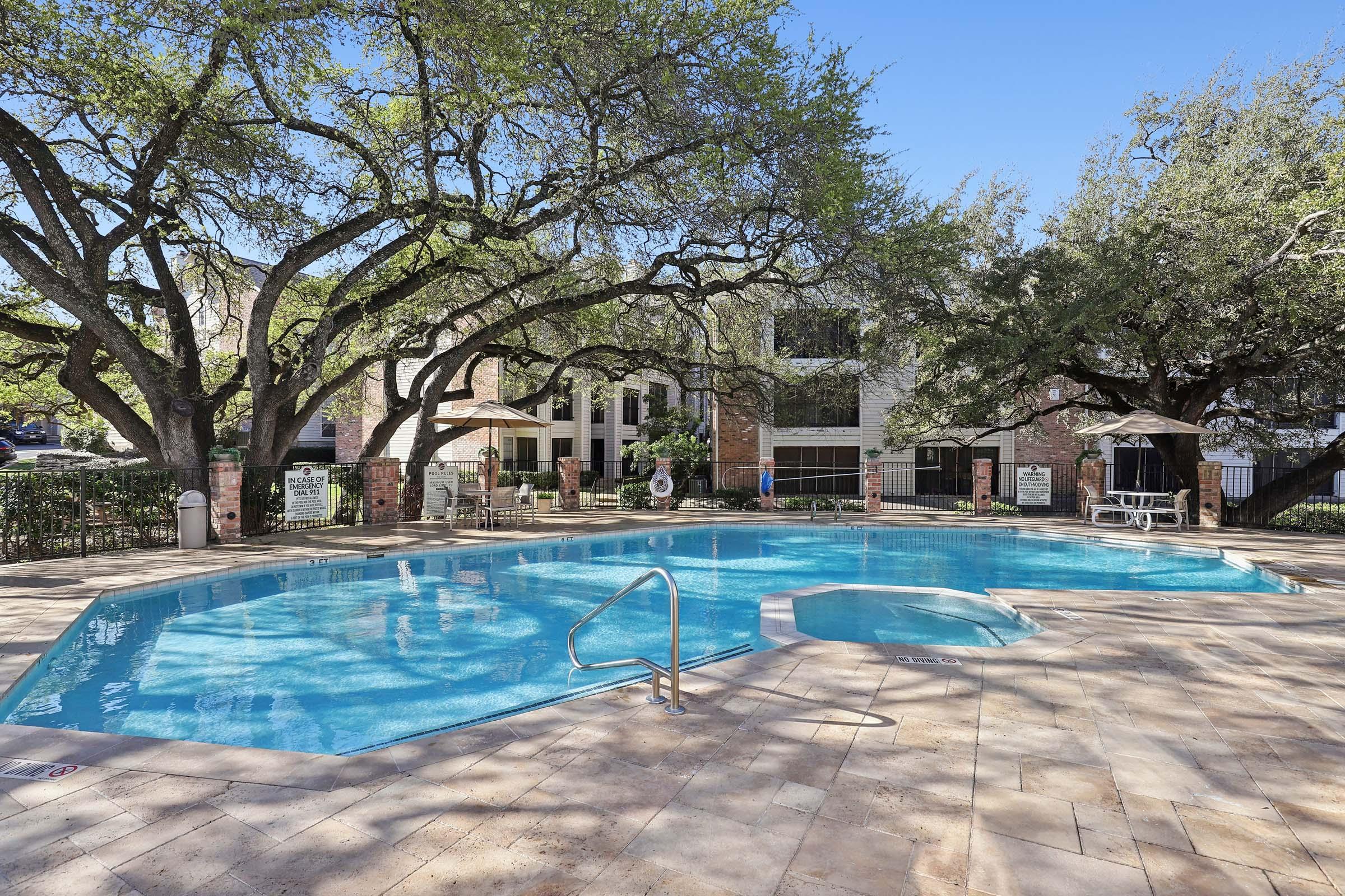 A clear blue swimming pool surrounded by lush green trees and a landscaped area. The pool features a hot tub adjacent to it, with lounge chairs and shaded seating areas nearby. The backdrop includes a residential building, creating a serene and inviting outdoor space.