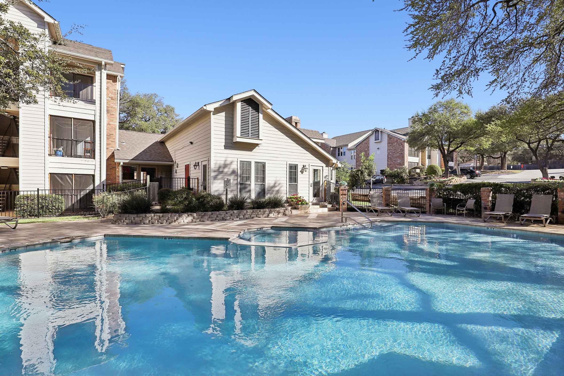 A clear blue swimming pool surrounded by lounge chairs and lush landscaping, with an apartment building visible in the background. The scene is set on a sunny day with a clear blue sky.