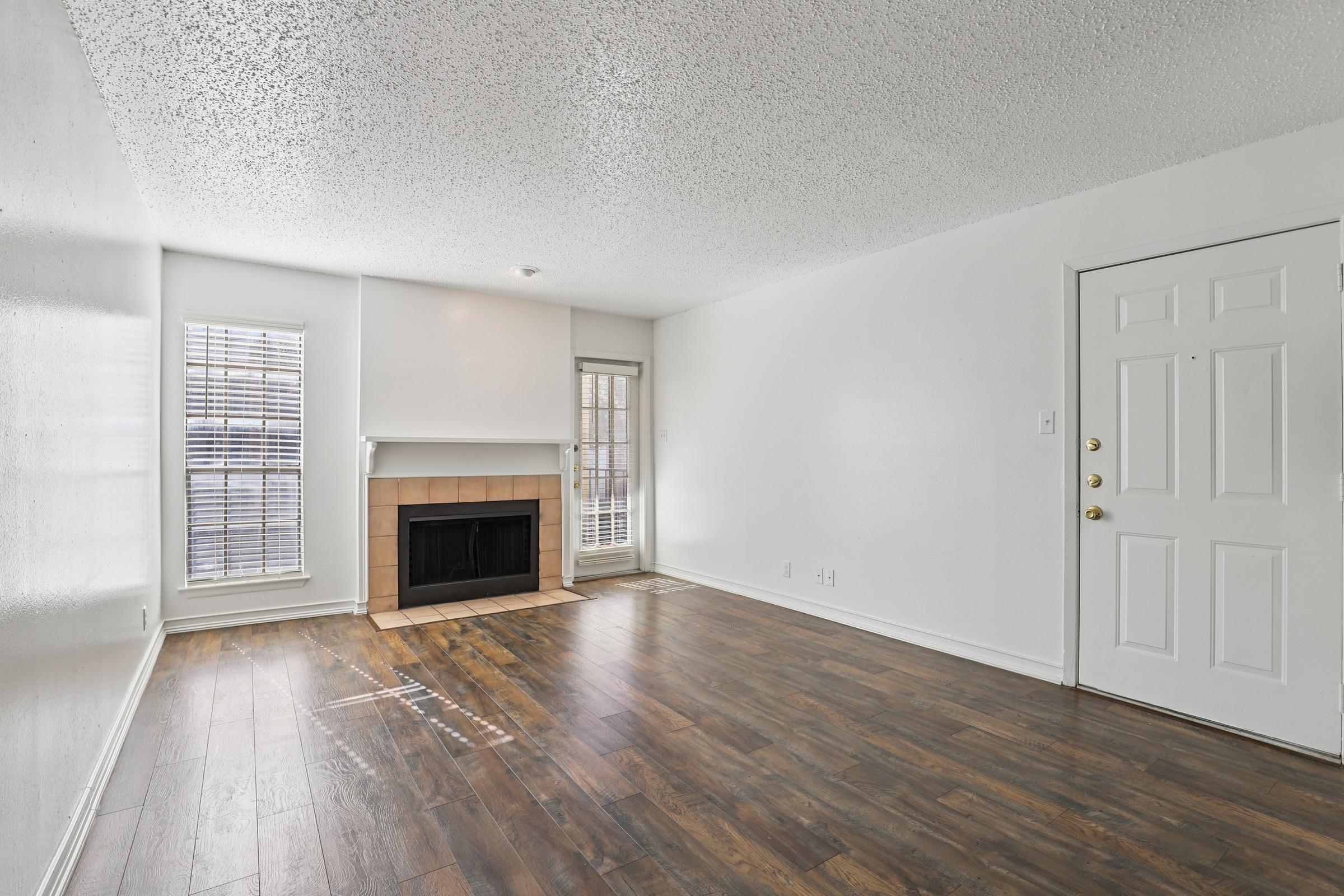 Spacious living room featuring a fireplace, large windows allowing natural light, and a front door. The floor is made of dark wood, and the walls are painted white, creating a bright and inviting atmosphere.