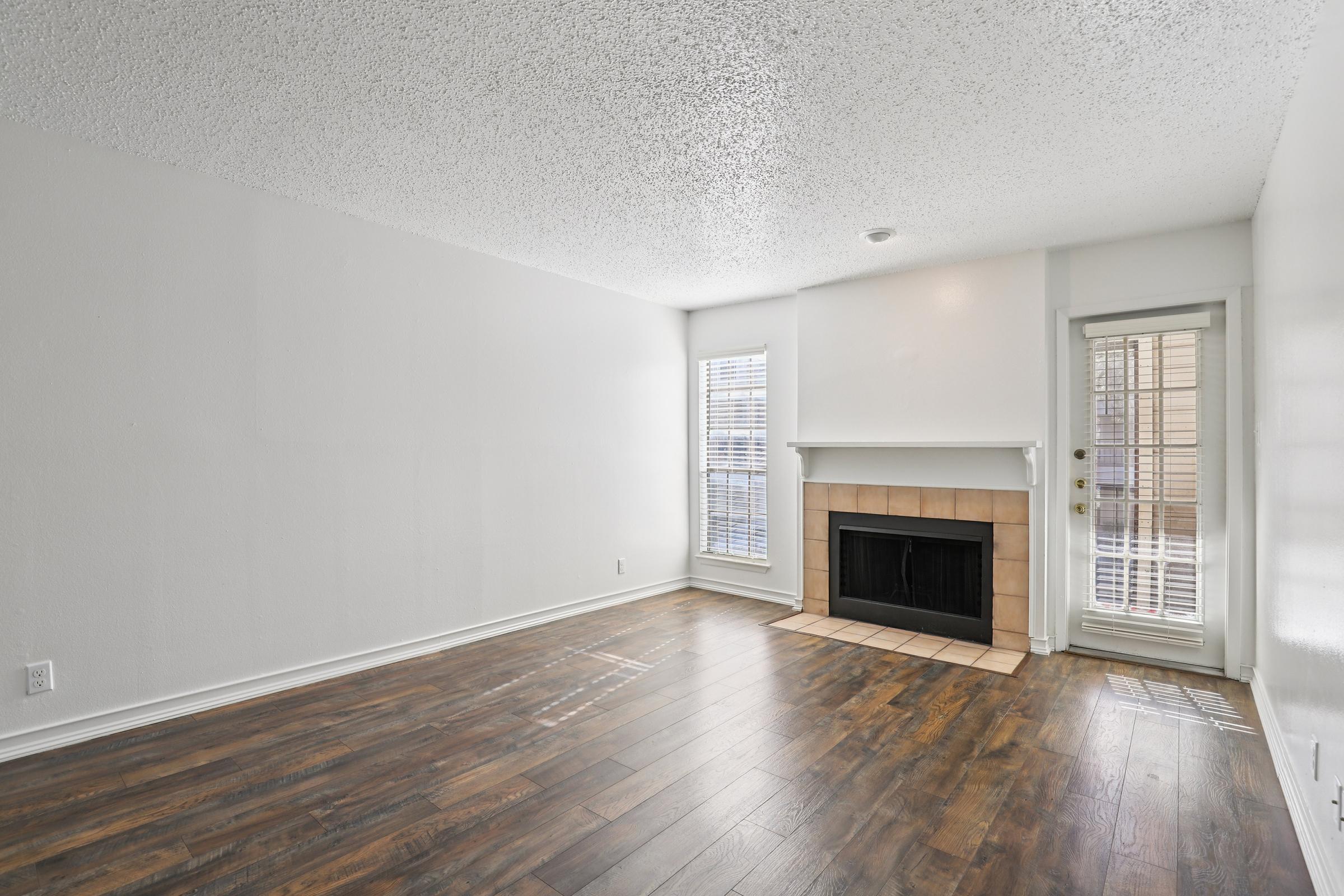 A spacious and empty living room featuring a textured ceiling, large windows allowing natural light, a white-painted wall, a fireplace with a tiled surround, and a door leading outside. The floor is covered in brown wooden planks, creating a warm and inviting atmosphere.