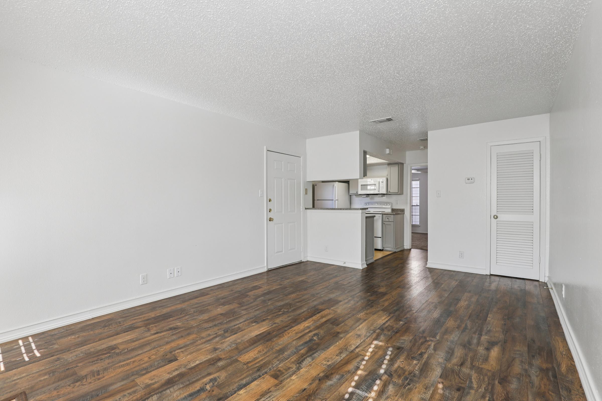 Interior view of a bright, empty living room with laminate flooring. A door leads outside, and there's a doorway to a kitchen area in the background. The walls are painted white, and there's a closet with louvered doors on the right. Natural light streams in, creating an open and inviting space.
