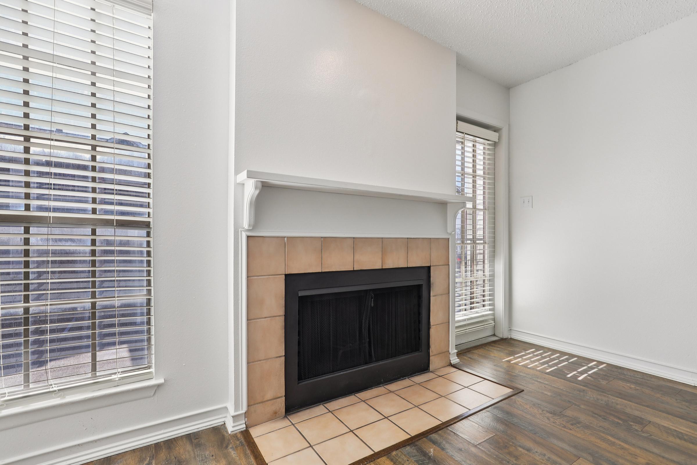A bright room featuring a white wall with a fireplace framed by beige tiles. Large windows with horizontal blinds allow natural light to fill the space, showcasing a wooden floor. The minimalist design creates a warm and inviting atmosphere.