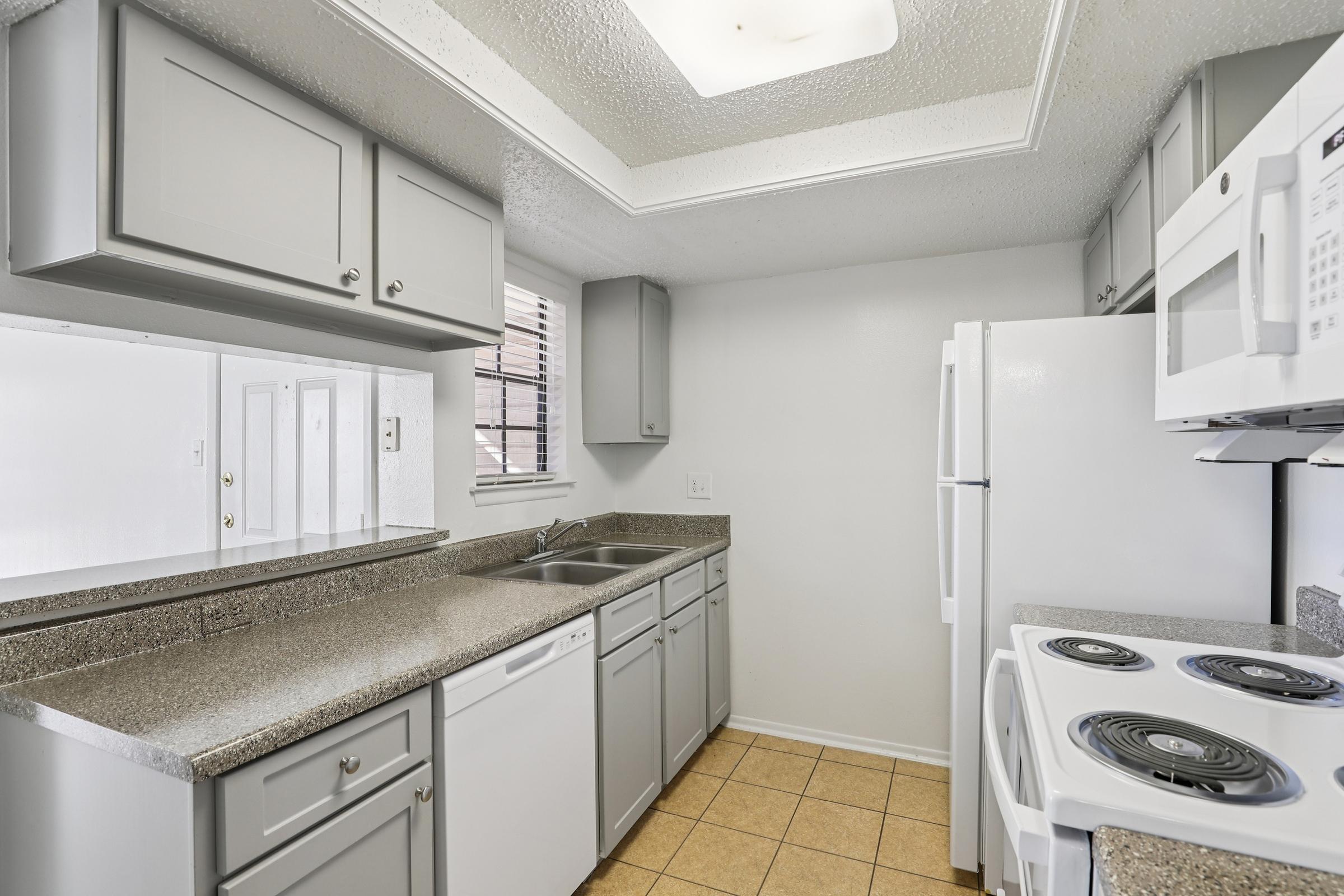 A modern kitchen featuring light gray cabinets, a double sink, and a beige countertop. Appliances include a white refrigerator, a microwave, and a white stove with a black cooktop. The room has tiled flooring and a window allowing natural light.