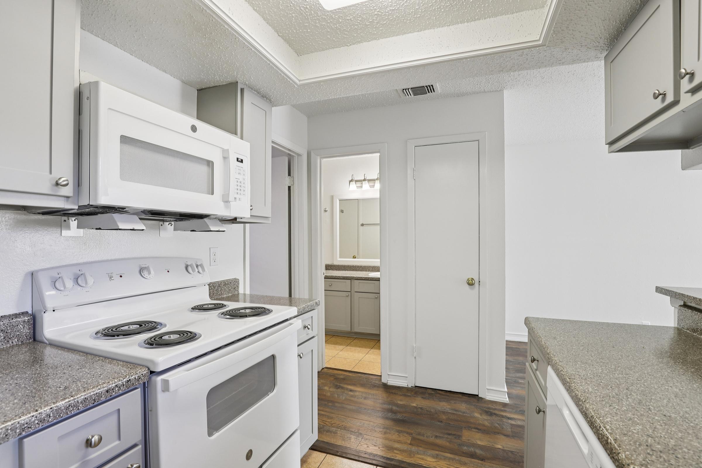 A modern kitchen featuring light gray cabinetry, a white stove and microwave, and granite countertops. In the background, a doorway leads to a bathroom with a mirror and vanity. The floor is a dark wood finish, adding contrast to the bright space.