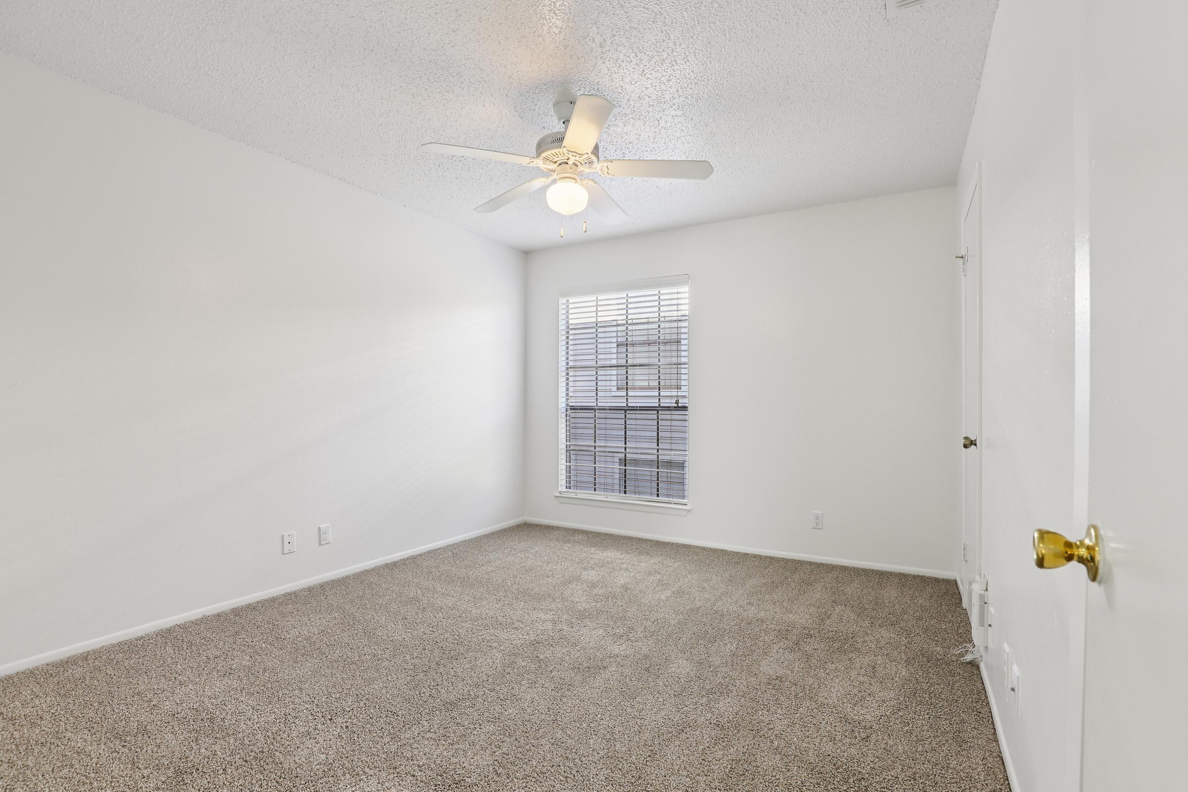 A bright, empty room with light-colored walls and a ceiling fan. The floor is covered in cozy beige carpeting. A window with blinds allows natural light to fill the space, and a closed door is visible on the right side. The overall atmosphere is clean and inviting, ideal for personal decor.