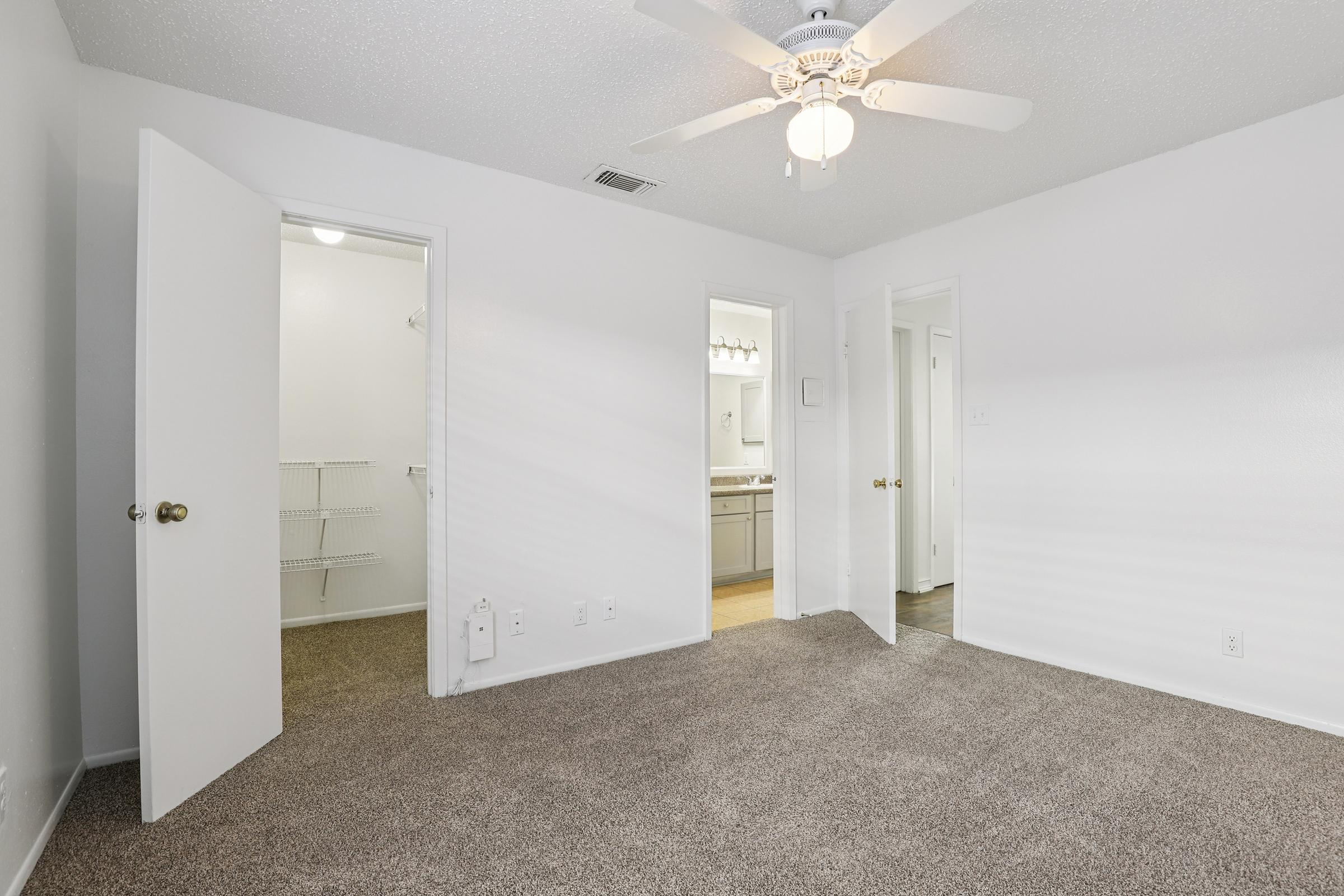 A spacious, well-lit bedroom featuring light-colored walls, a ceiling fan, and plush carpeting. Two open doors lead to additional rooms: one appears to be a bathroom and the other a closet. The overall design is simple and inviting, ideal for a comfortable living space.