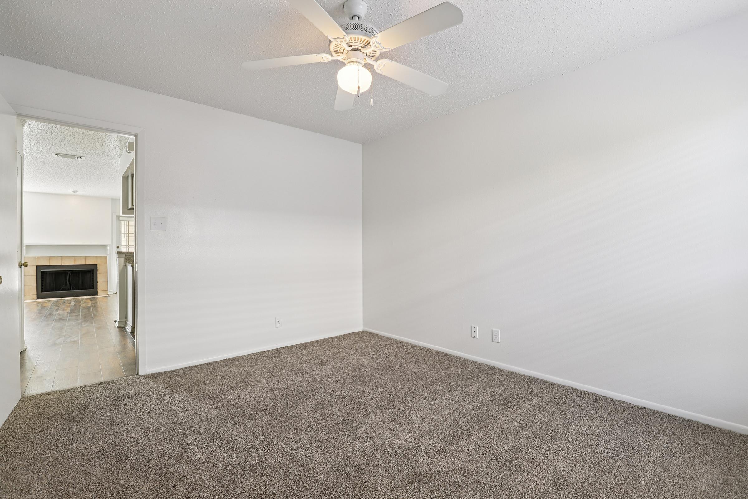 Empty room with light beige carpet and a ceiling fan. A door leads to another room, and a fireplace is visible in the distance. The walls are painted white, creating a bright and airy atmosphere. Natural light streams in, enhancing the spacious feel of the room.