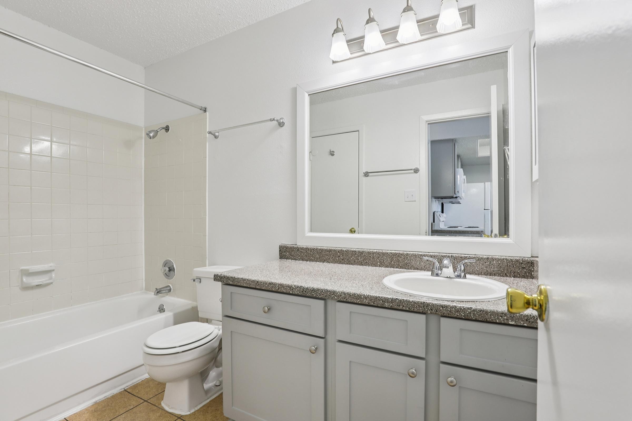 A clean and modern bathroom featuring a bathtub with a shower, a white toilet, and a double sink vanity with gray cabinets. The walls are painted white, and there is a large mirror above the sink. Soft lighting is provided by four wall-mounted fixtures. A door leads to another room in the background.