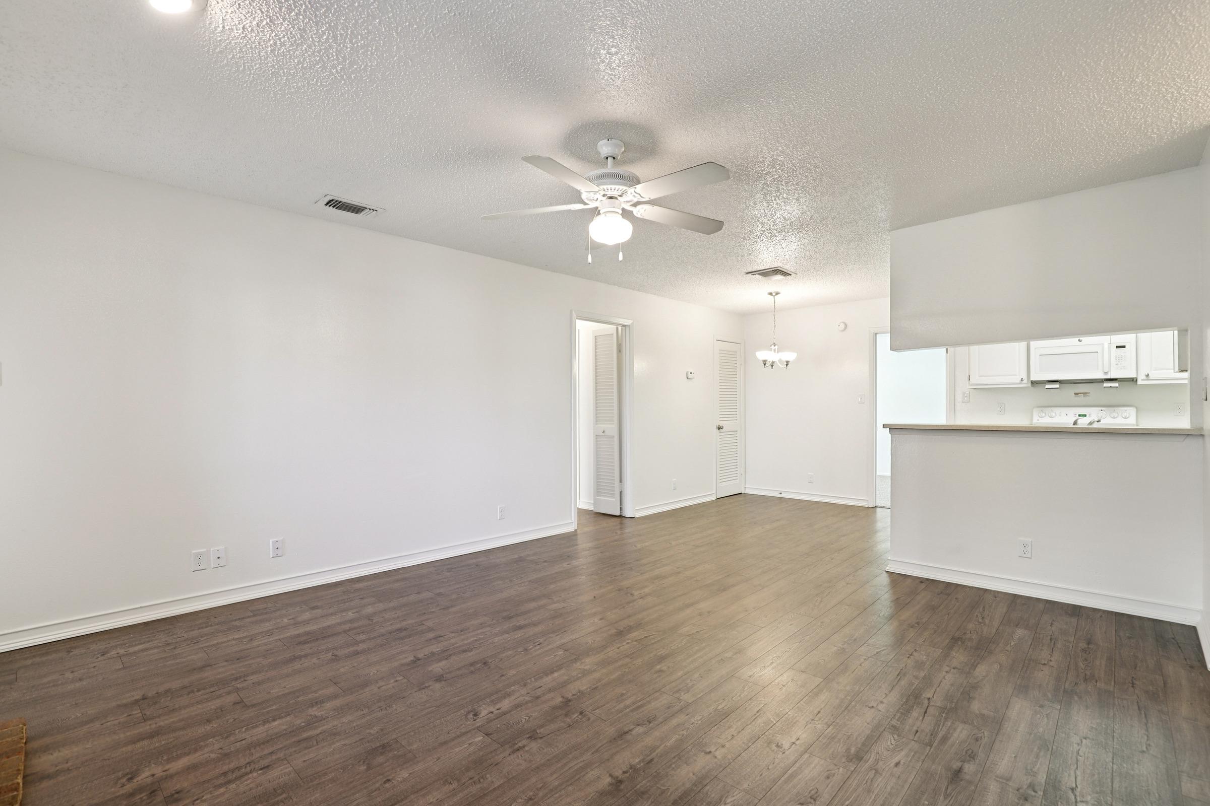 A spacious, well-lit living room featuring a ceiling fan, light-colored walls, and dark laminate flooring. A doorway leads to a small hallway, while an adjacent area reveals a kitchen with white cabinetry. The open layout enhances the room’s airy feel.